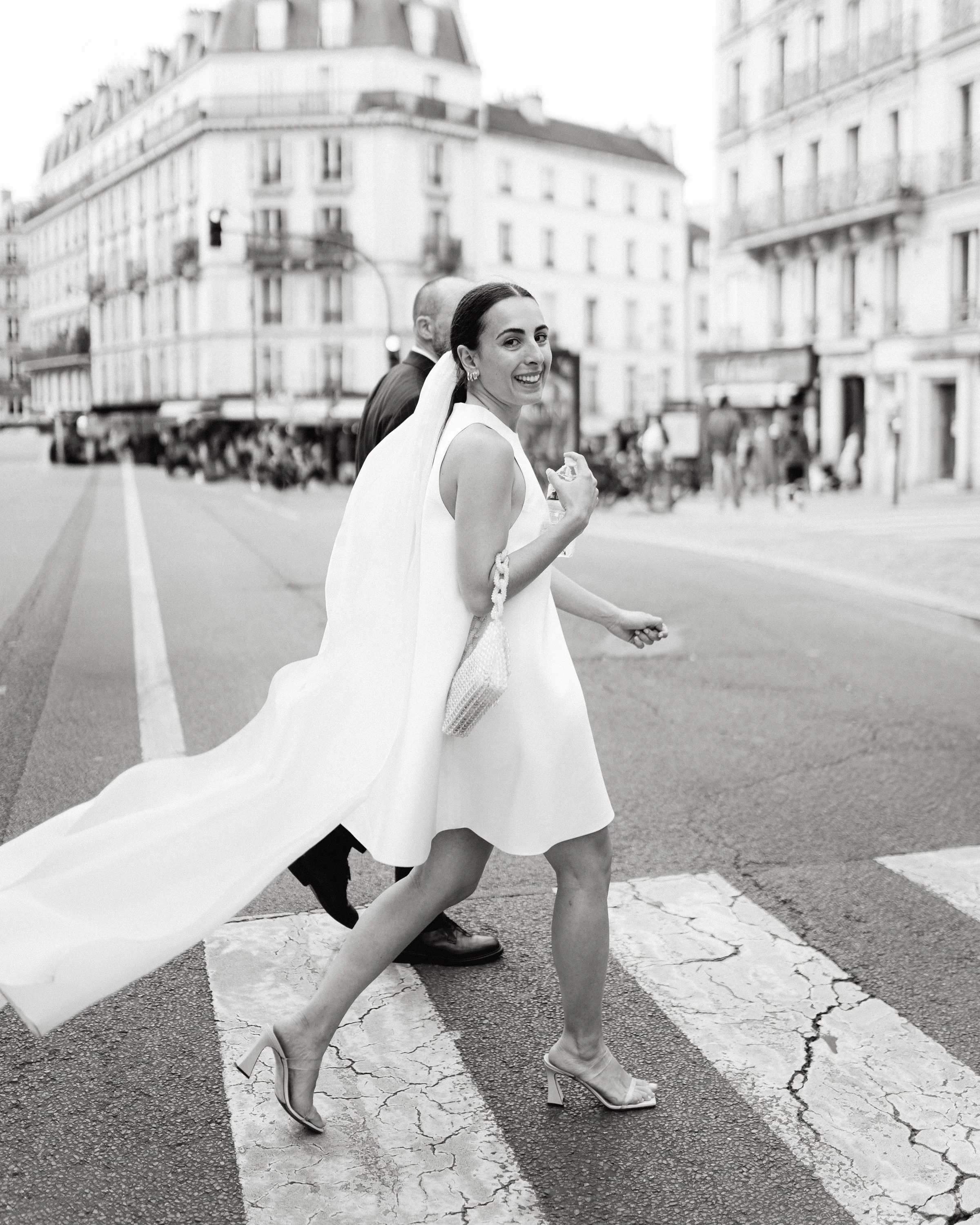 Une femme souriante, habillée en robe blanche de style élégant, portant des talons hauts et un peignoir, traverse un passage piéton dans une rue urbaine, avec des bâtiments anciens en arrière-plan.