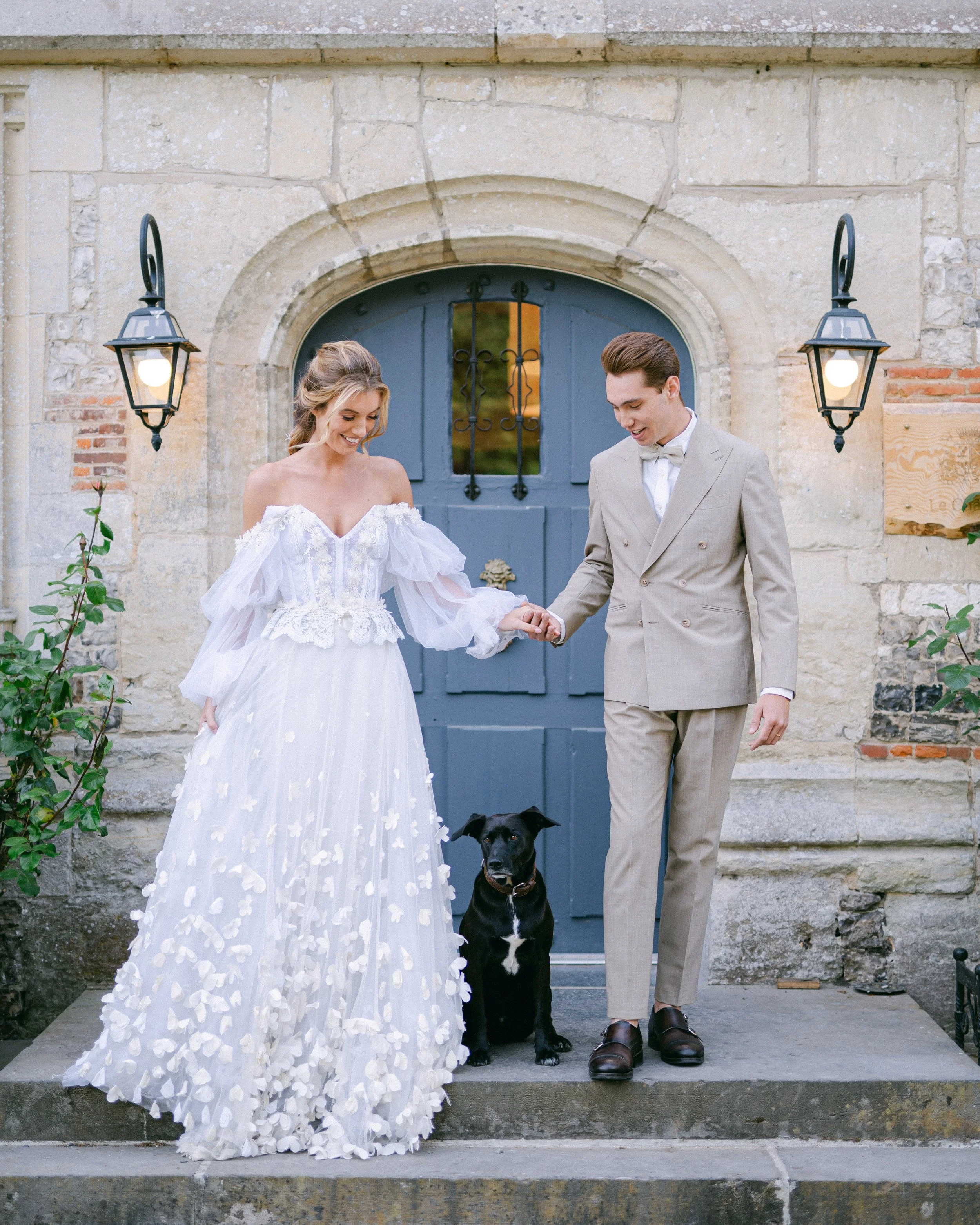 Un couple en mariage, tenant un chien, devant une porte bleue ancienne, dans une façade en pierre, lors d'une journée ensoleillée.