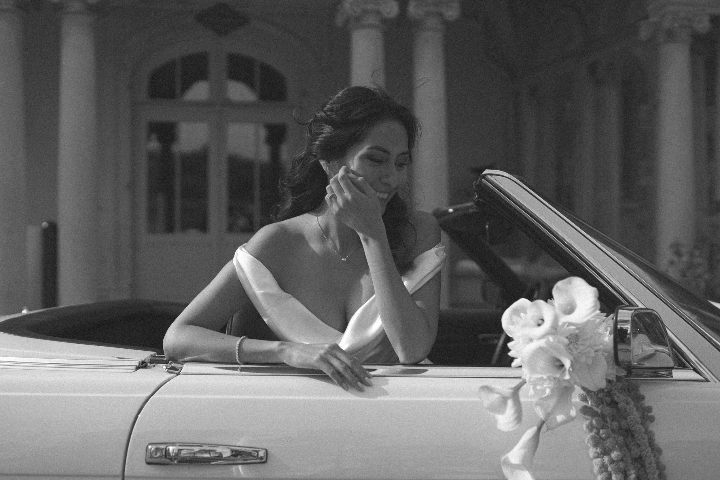 Une femme souriante sortant d'une voiture décapotable, décorée de fleurs, lors d'une occasion spéciale, en noir et blanc.