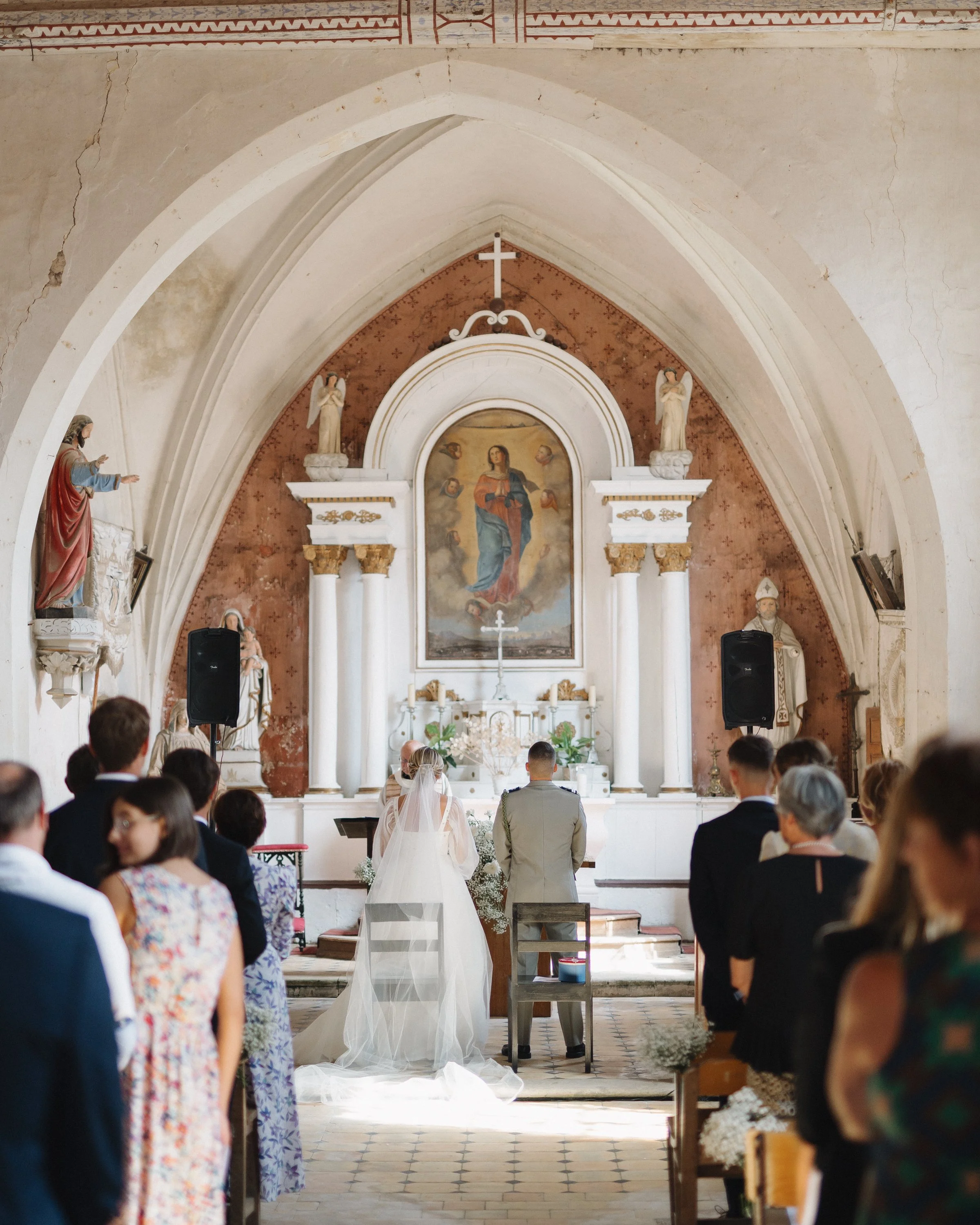 Cérémonie de mariage dans une église ancienne avec des invités assis, une mariée en robe blanche et un homme en uniforme, devant un autel avec une peinture de la Vierge Marie, entourés de statues et colonnes. 