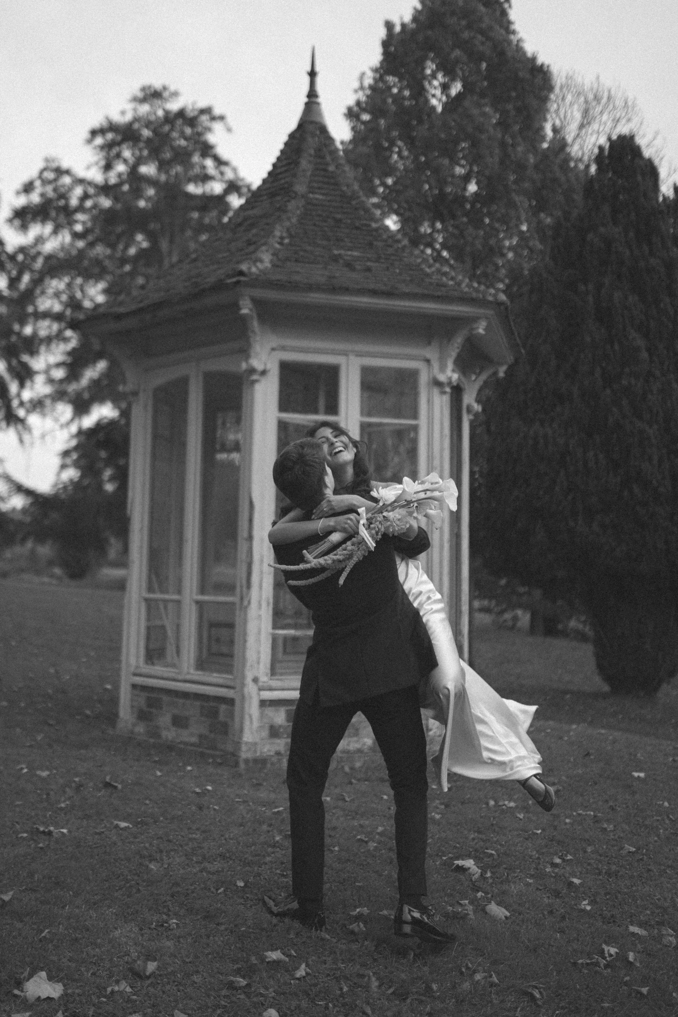 Un couple en habits de mariage, l'homme levant la femme en l'air, riant, devant une petite maison en bois dans un parc, en noir et blanc.