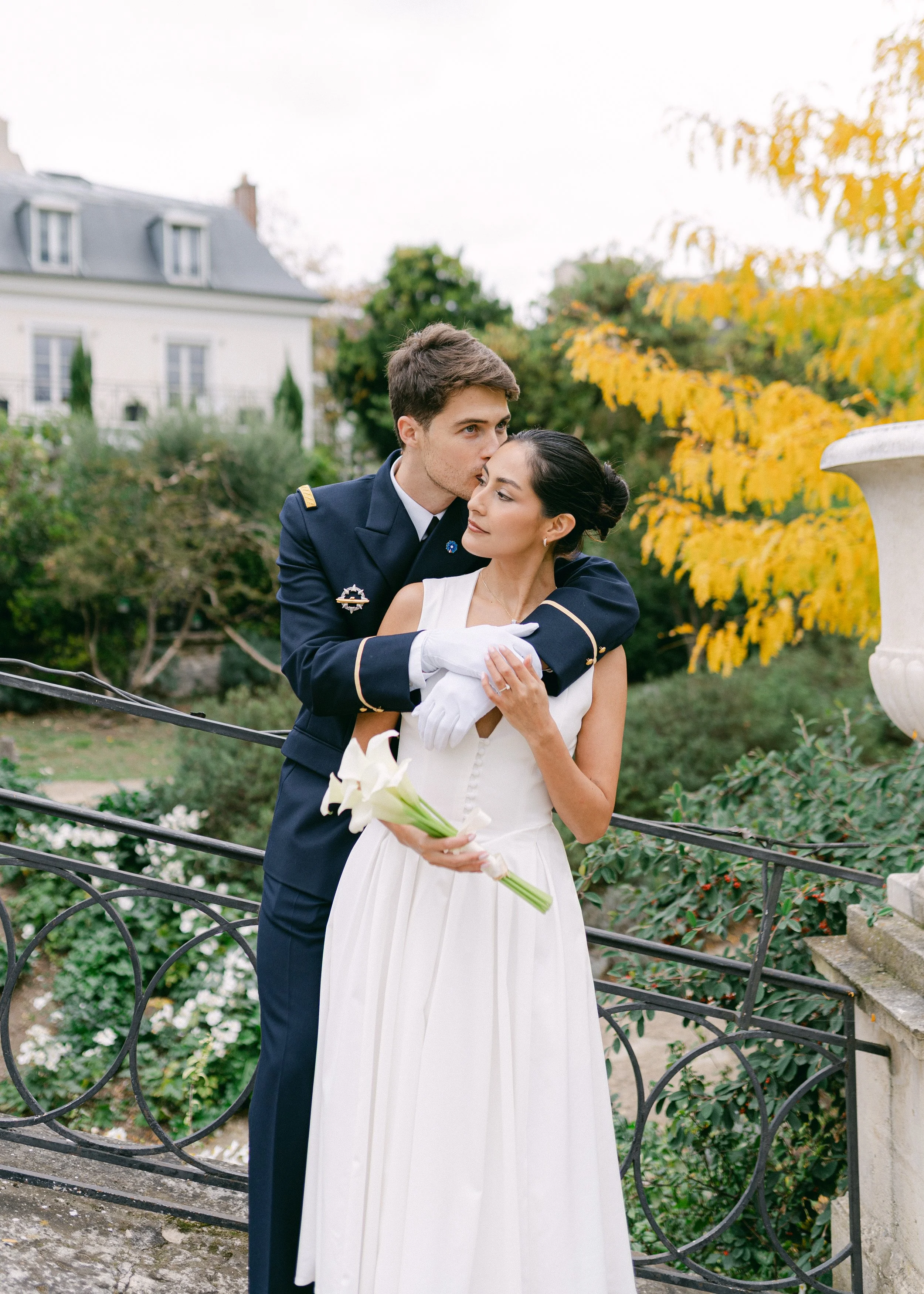Un couple en tenue de mariage à l'extérieur, avec un homme en uniforme militaire et une femme en robe blanche, la femme tient un bouquet de calla lilies.