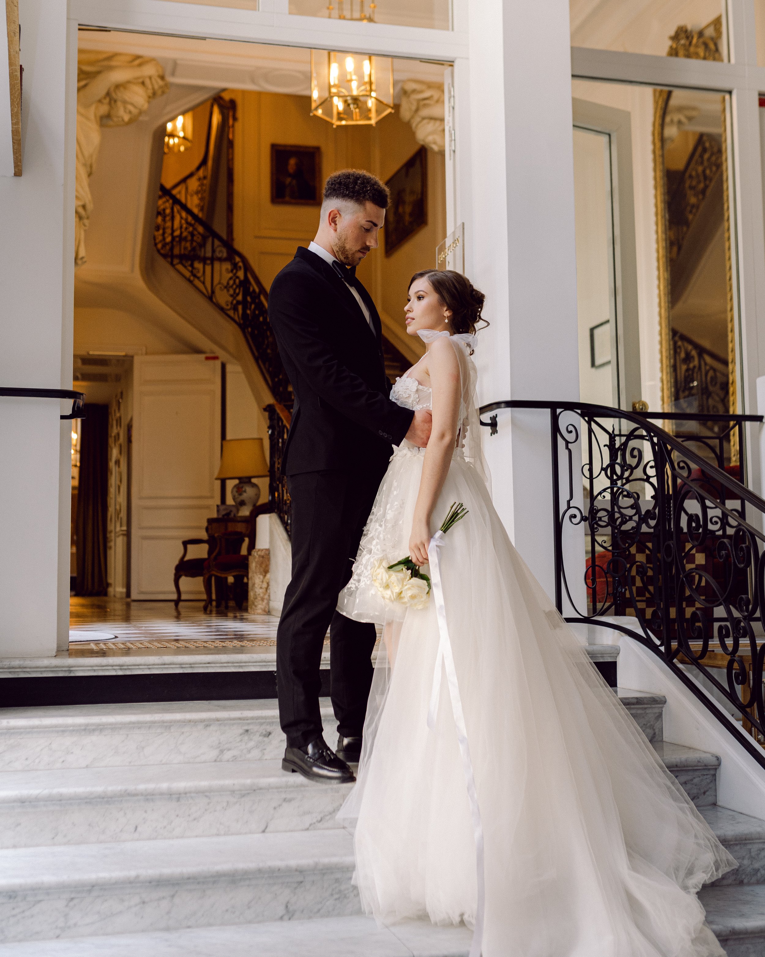 Un couple de mariés posant sur un escalier en marbre dans un intérieur élégant, la femme en robe de mariage blanche tenant un bouquet et le homme en costume noir.