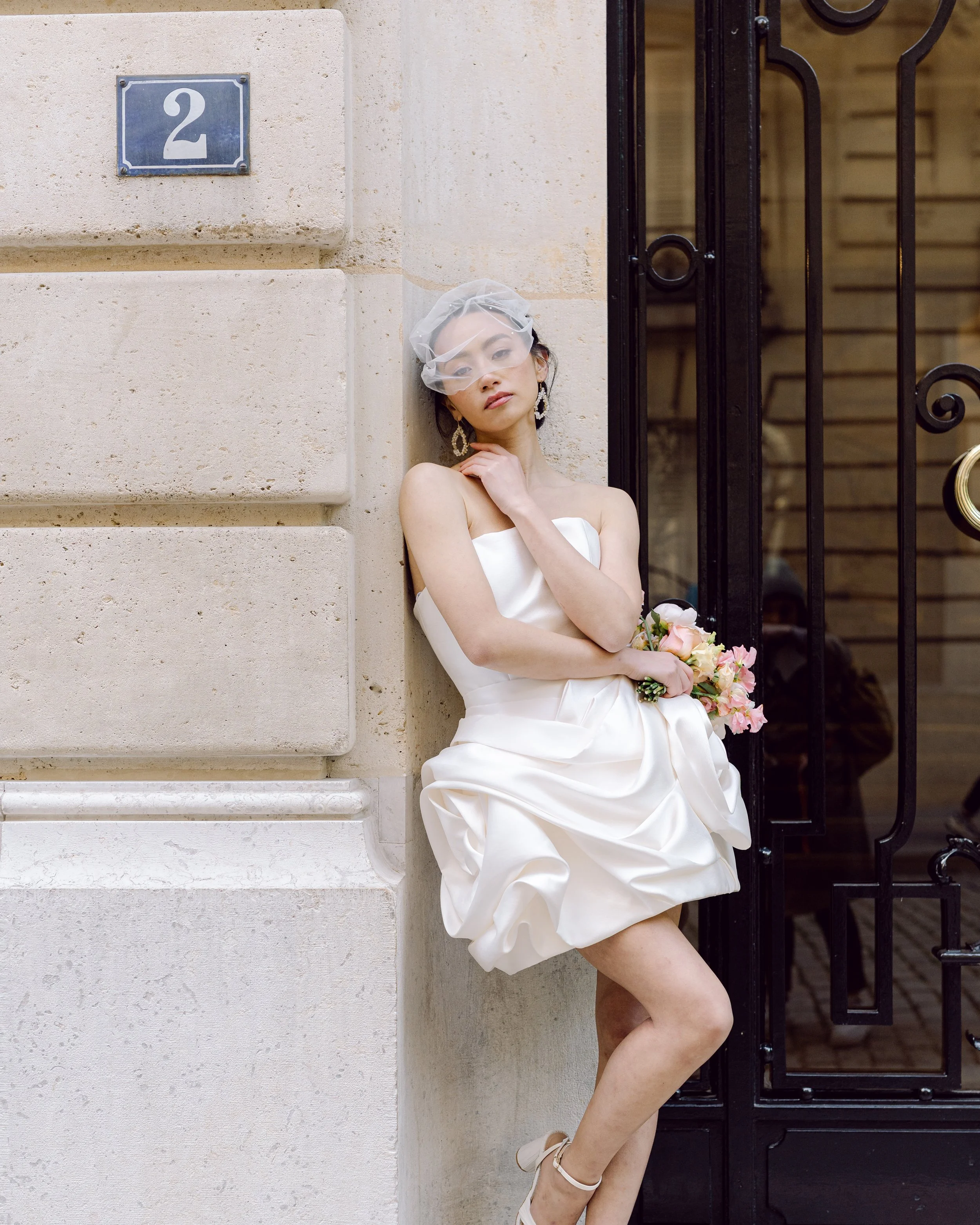 Une femme portant une robe de mariage blanche, tenant un bouquet de fleurs roses, posant contre un mur en pierre à côté d'une porte en fer noire.