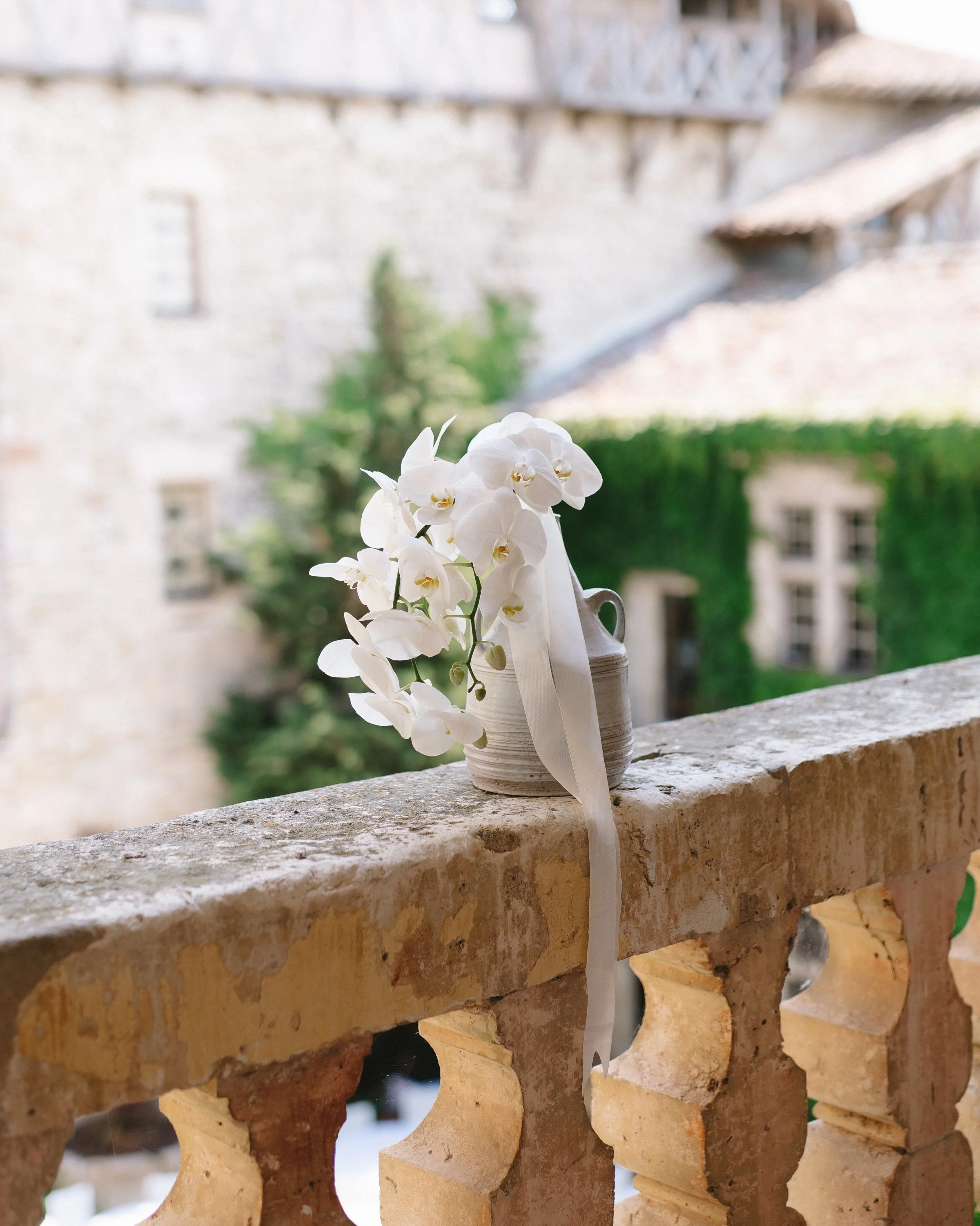Bouquet de fleurs blanches dans un vase posé sur un balcon en pierre, avec un bâtiment ancien en pierre en arrière-plan.