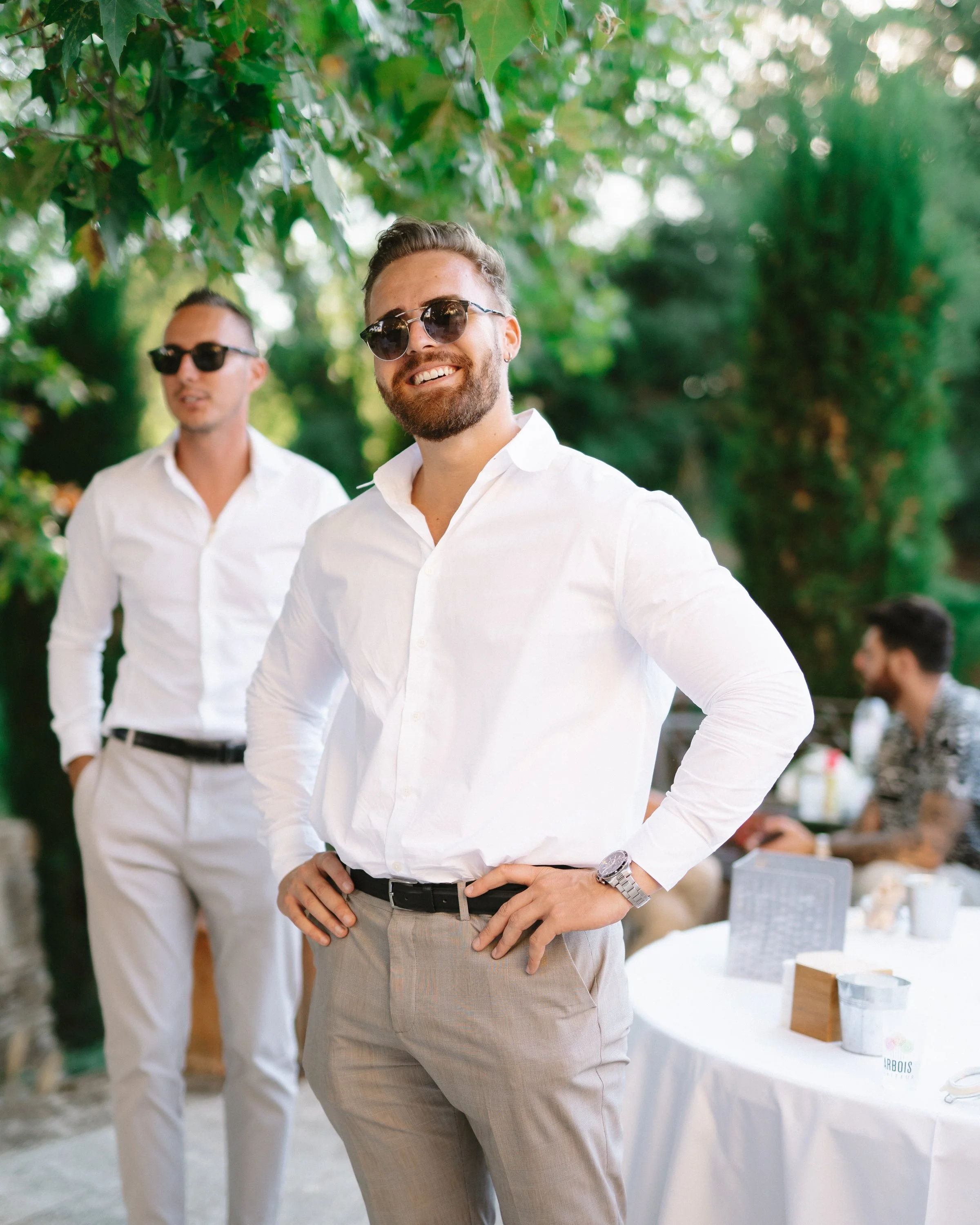 Deux hommes souriants portant des chemises blanches et des lunettes de soleil, se tiennent en extérieur sous un arbre, dans un environnement décontracté et ensoleillé.