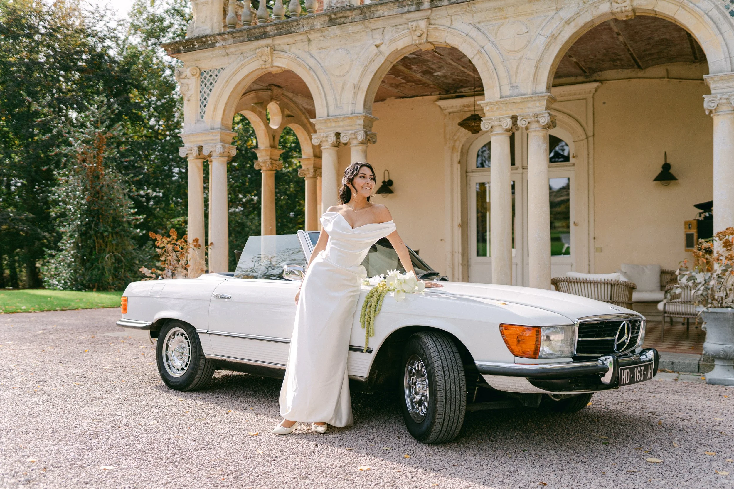 Une femme en robe de mariage posant à côté d'une voiture blanche Mercedes-Benz devant une maison ancienne avec colonnes, dans un cadre extérieur avec des arbres.