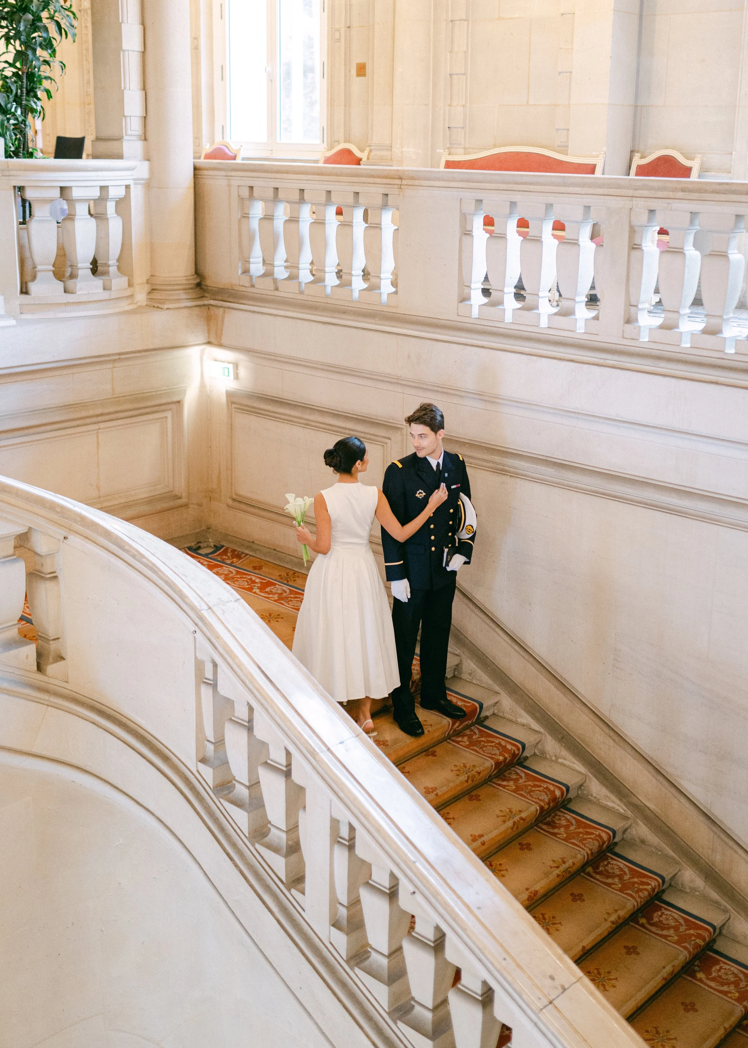 Une femme en robe blanche offre des fleurs à un homme en uniforme militaire dans un bâtiment élégant avec des escaliers en marbre.