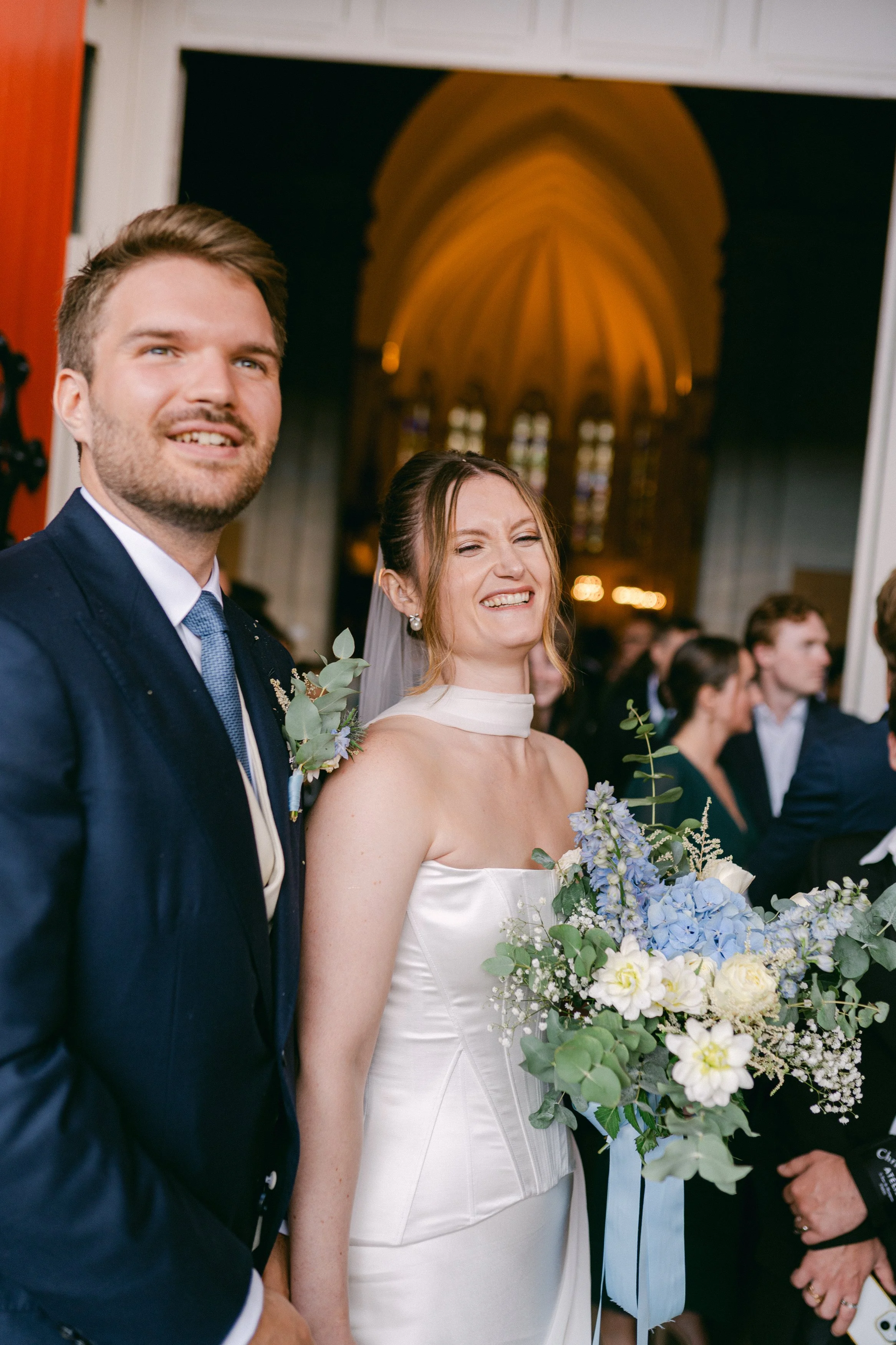 Un couple lors d'un mariage, la femme avec une robe blanche et le homme en costume, tenant un bouquet de fleurs blanches et bleues, dans une église.