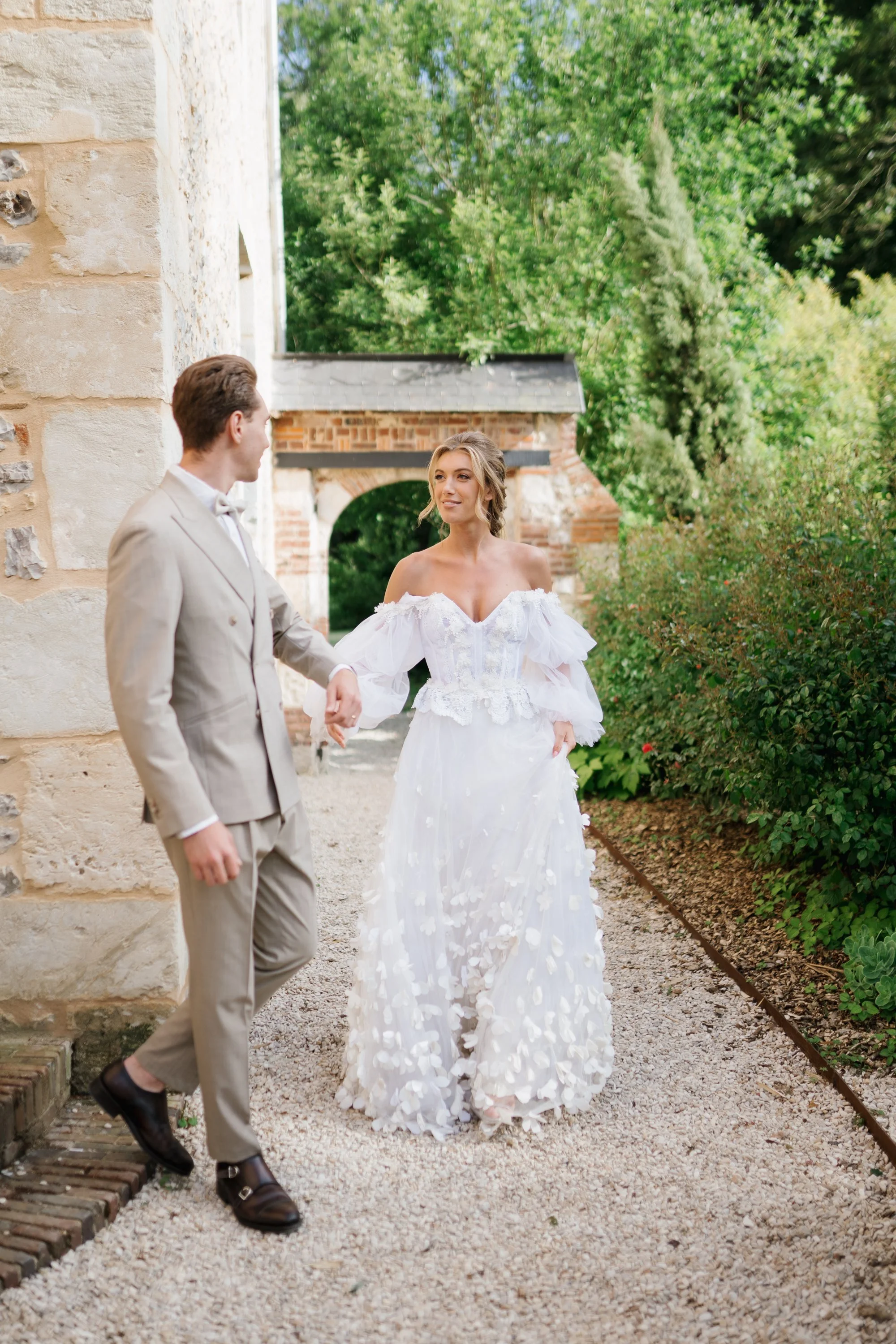Un couple en mariage, la femme en robe blanche longue avec des détails floraux et l'homme en costume beige, se tiennent dans un jardin verdoyant, en train de se tenir la main.