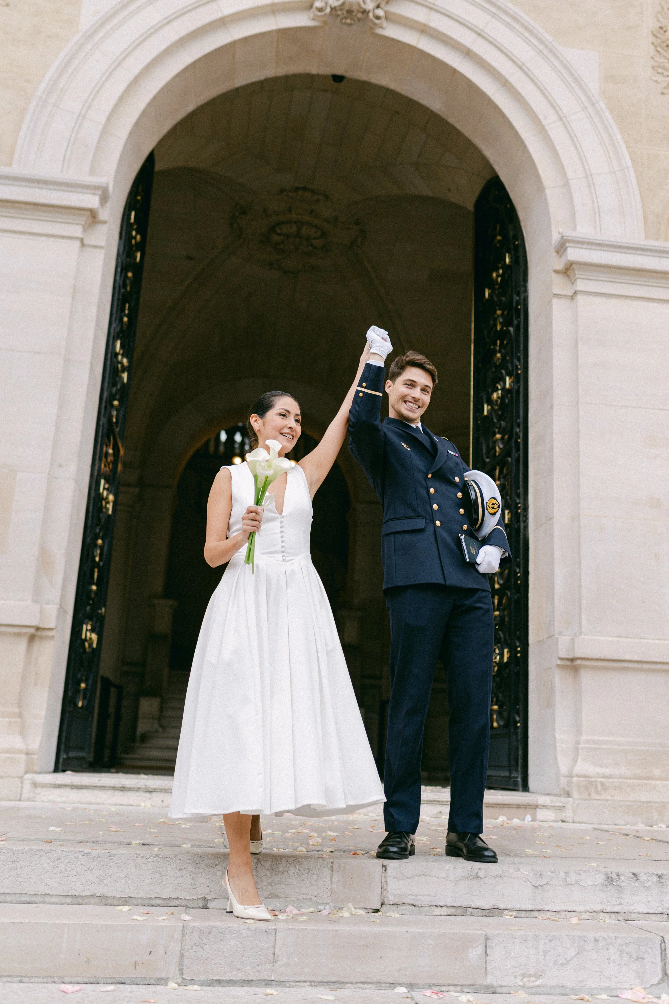 Un couple de mariés souriants se tenant la main en haut des marches, devant une entrée en pierre d'un bâtiment historique, la femme tient un bouquet de callas et porte une robe blanche, l'homme en uniforme militaire tient un casque.