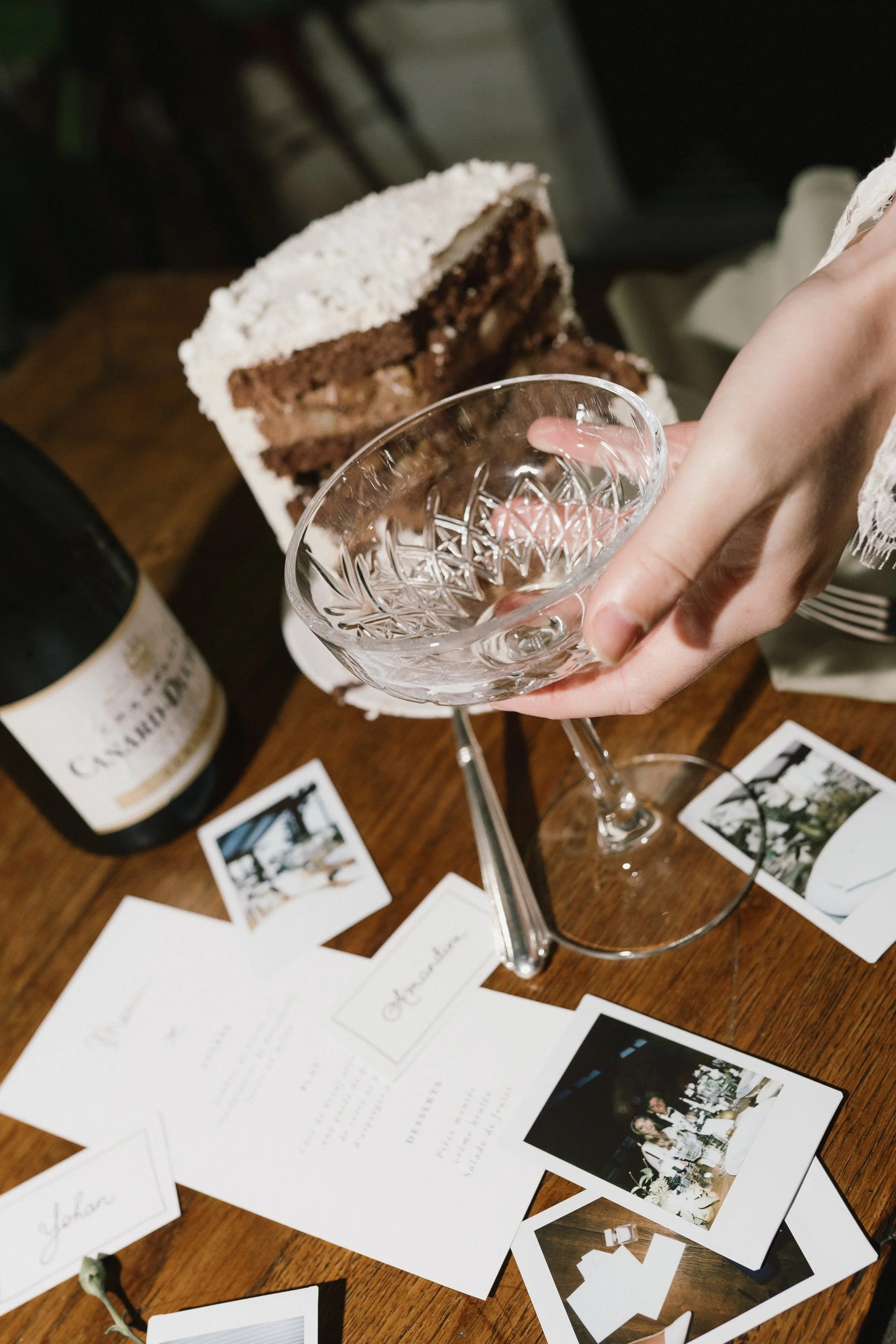 Une main tient un verre à cocktail vide, un gâteau au chocolat au fond, une bouteille de vin et des photos sur une table en bois.