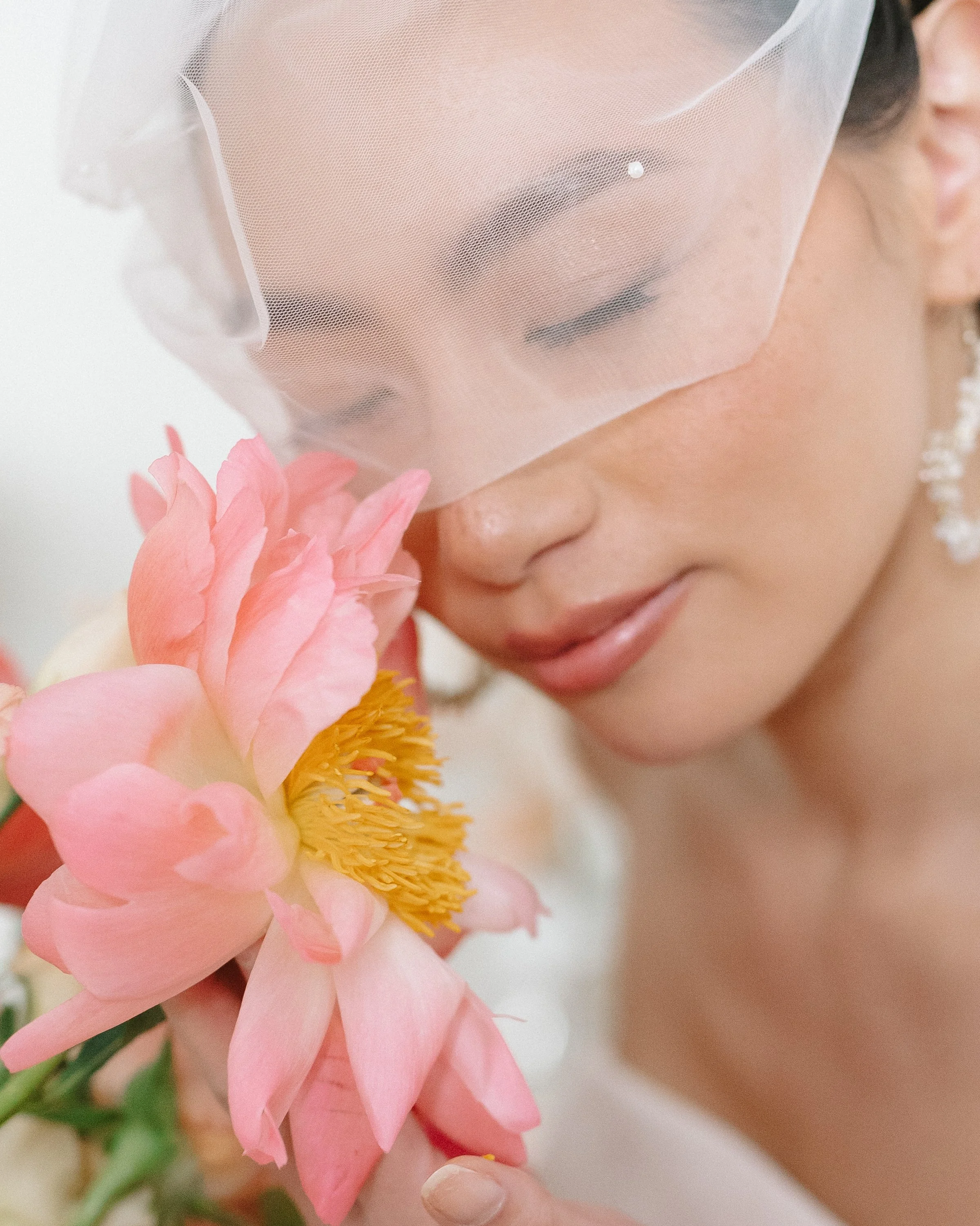 Une femme porte un voile de mariée, tient un bouquet de fleurs roses et regarde délicatement une grande fleur. Elle a des cheveux bruns et un maquillage subtil.