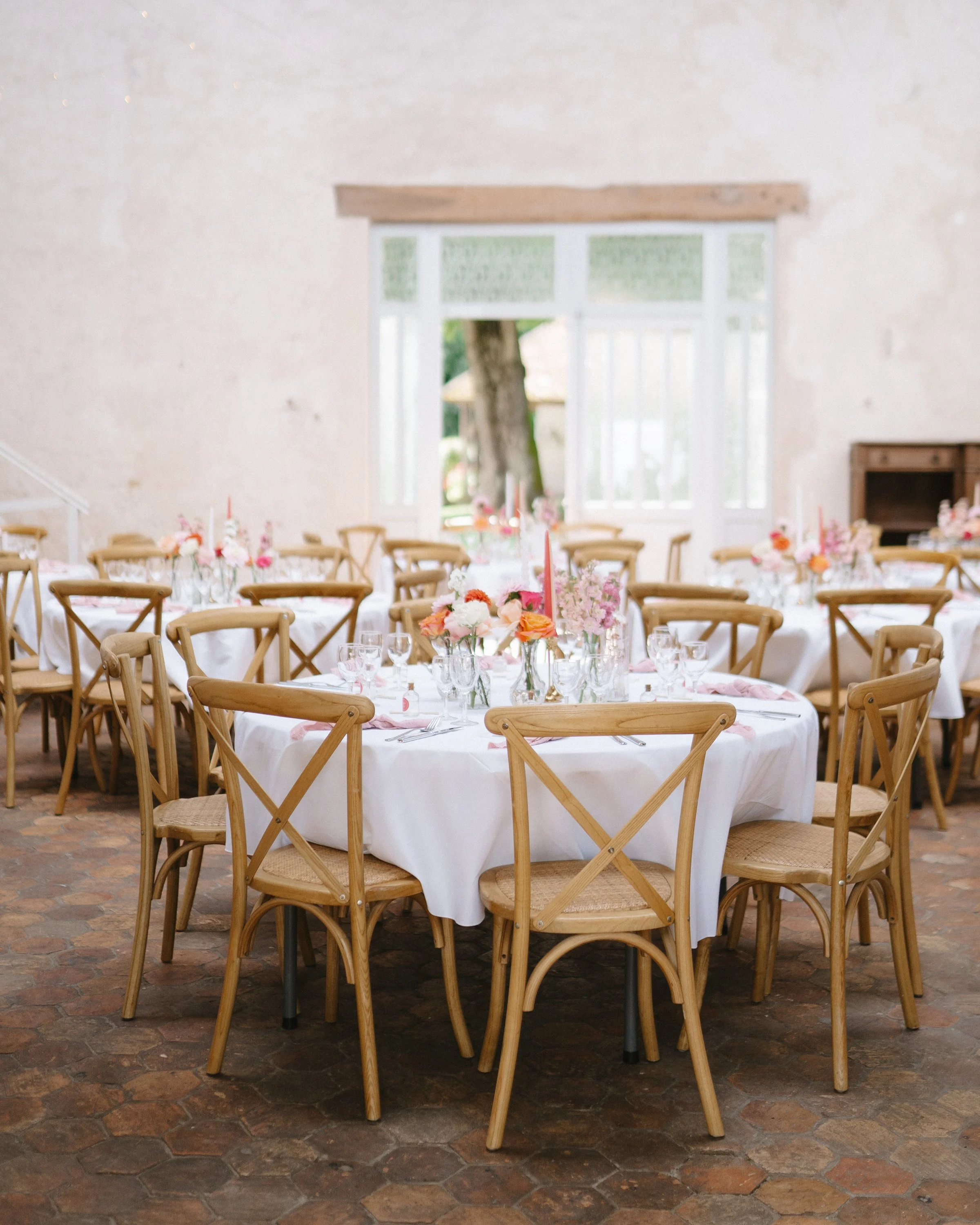 Salle de réception avec tables rondes, nappes blanches, décor floral pastel, chaises en bois, grande fenêtre laissant entrer la lumière naturelle.
