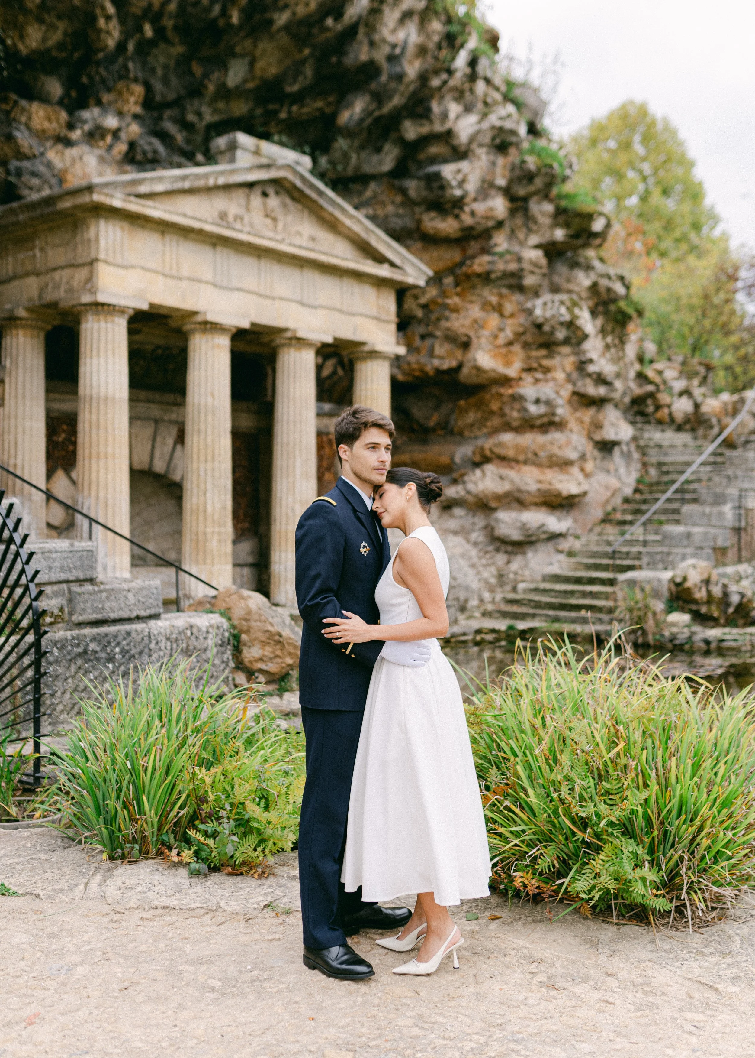 Un couple en habits élégants, un homme en uniforme militaire et une femme en robe blanche, se tiennent dans un jardin avec une ancienne structure en ruines en arrière-plan.