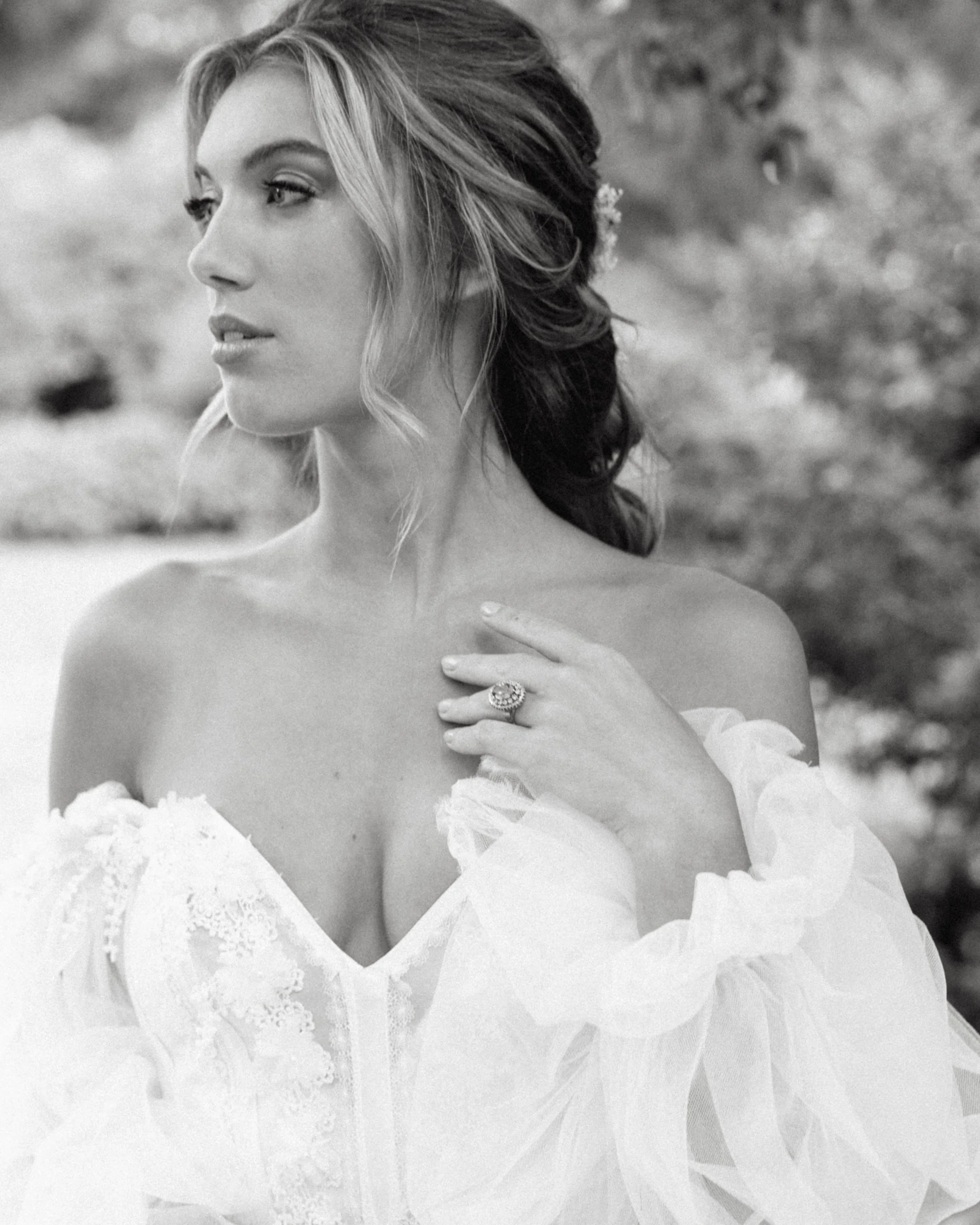 Une femme portant une robe de mariée en dentelle, avec des manches en tulle, se tenant à l'extérieur avec un arrière-plan flou de nature, et qui regarde vers la gauche, réalisant une séance photo nuptiale.