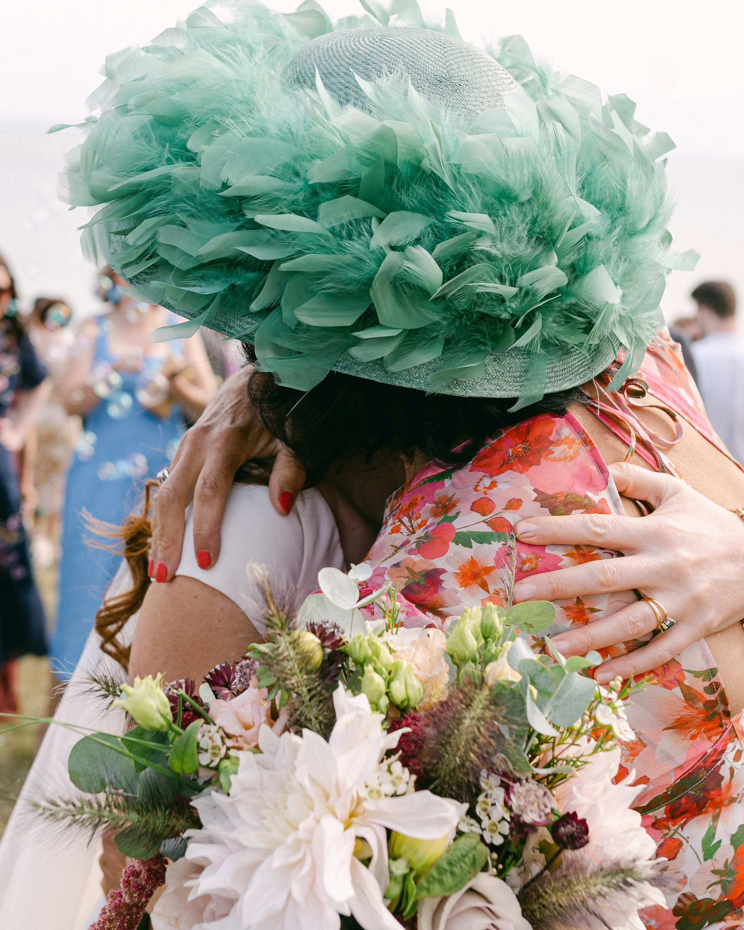 Deux personnes s'embrassent en se serrant dans les bras lors d'une célébration, portant des vêtements colorés et un chapeau décoré de plumes vertes, avec un bouquet de fleurs.