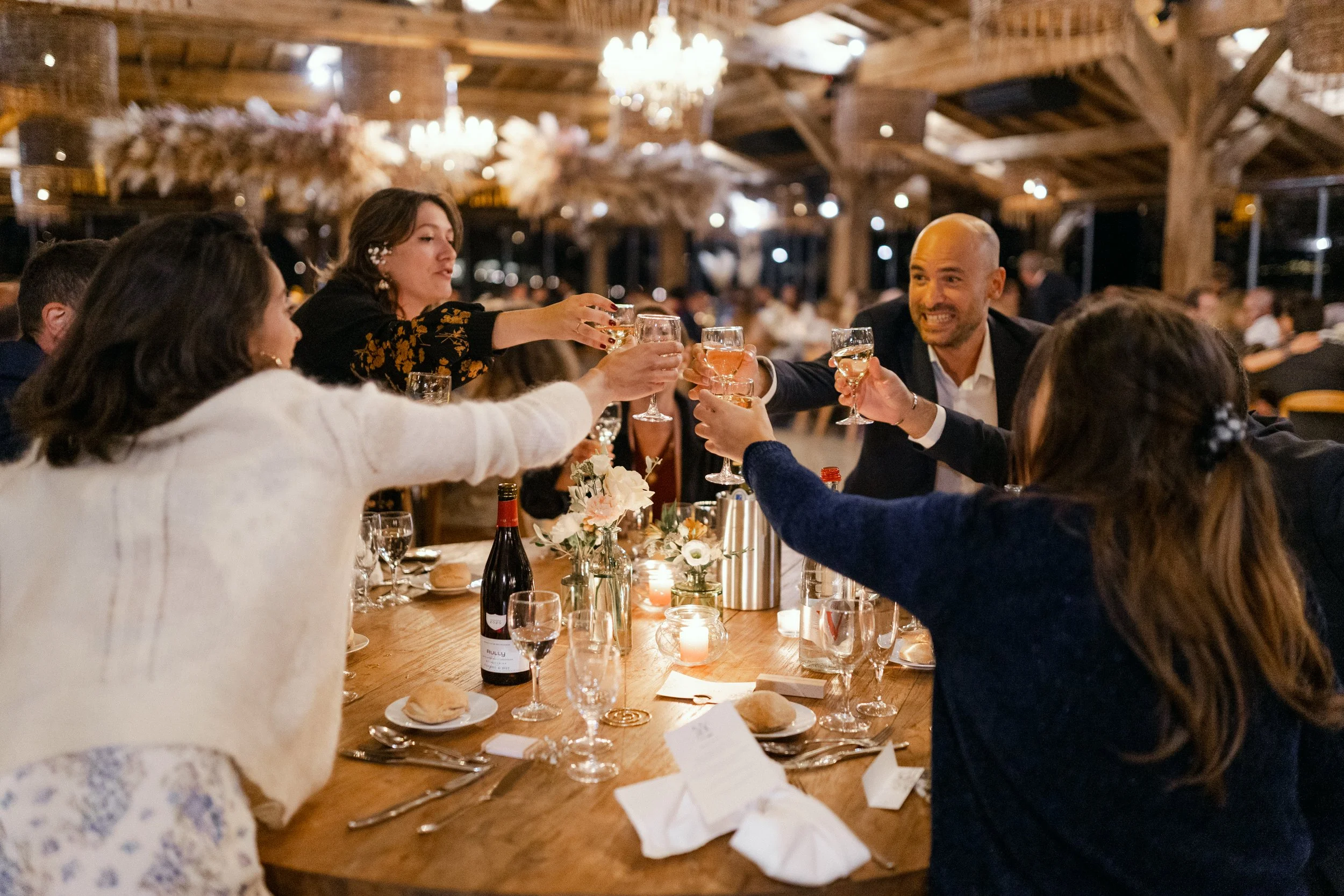 Groupe de personnes à une fête, levant des verres pour porter un toast dans un décor chaleureux avec lumières et chandeliers, assis autour d'une table en bois avec des fleurs et des bougies.