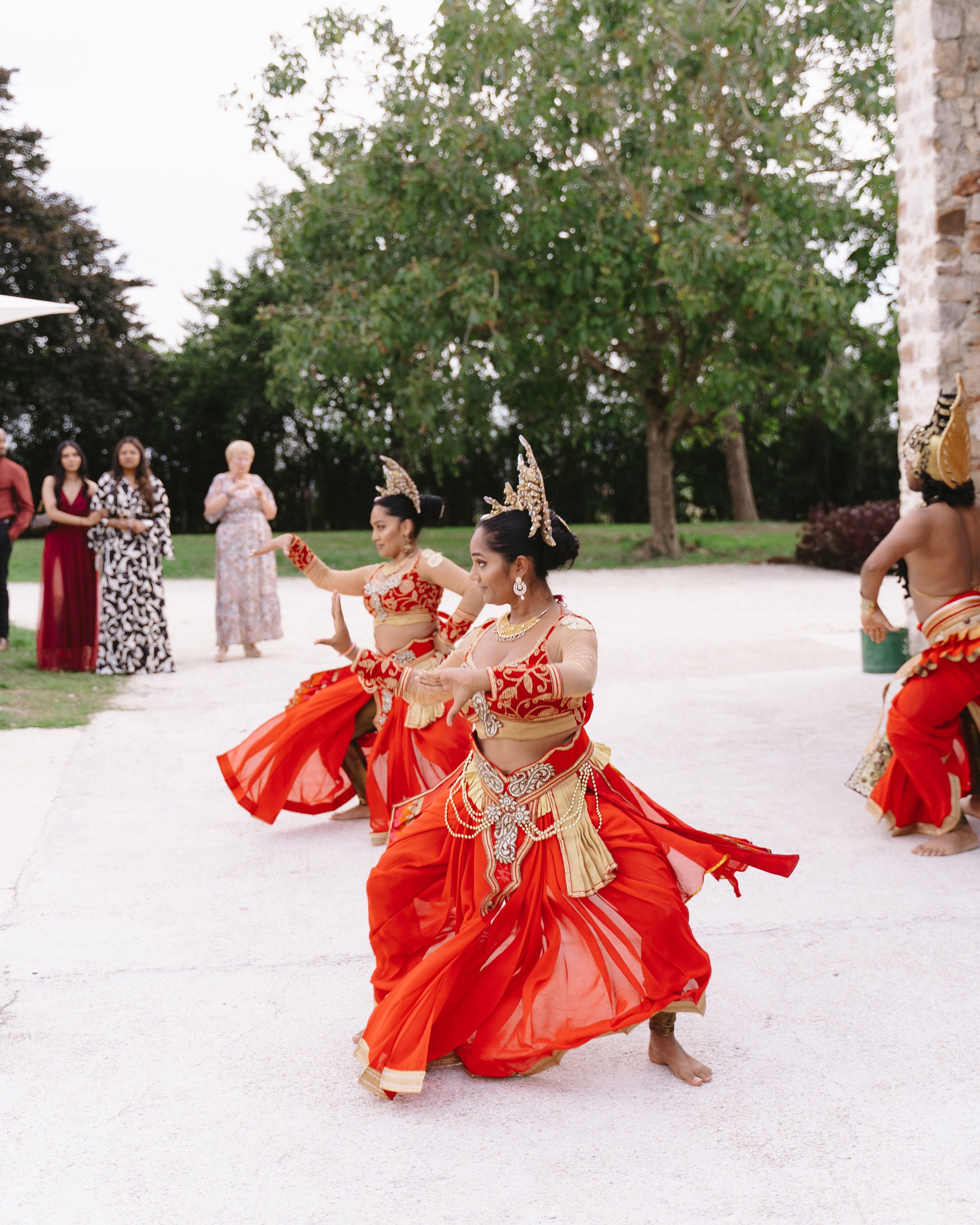 Trois femmes indiennes dansent en costumes traditionnels rouges, accompagnées d'autres personnes qui regardent, à l'extérieur.