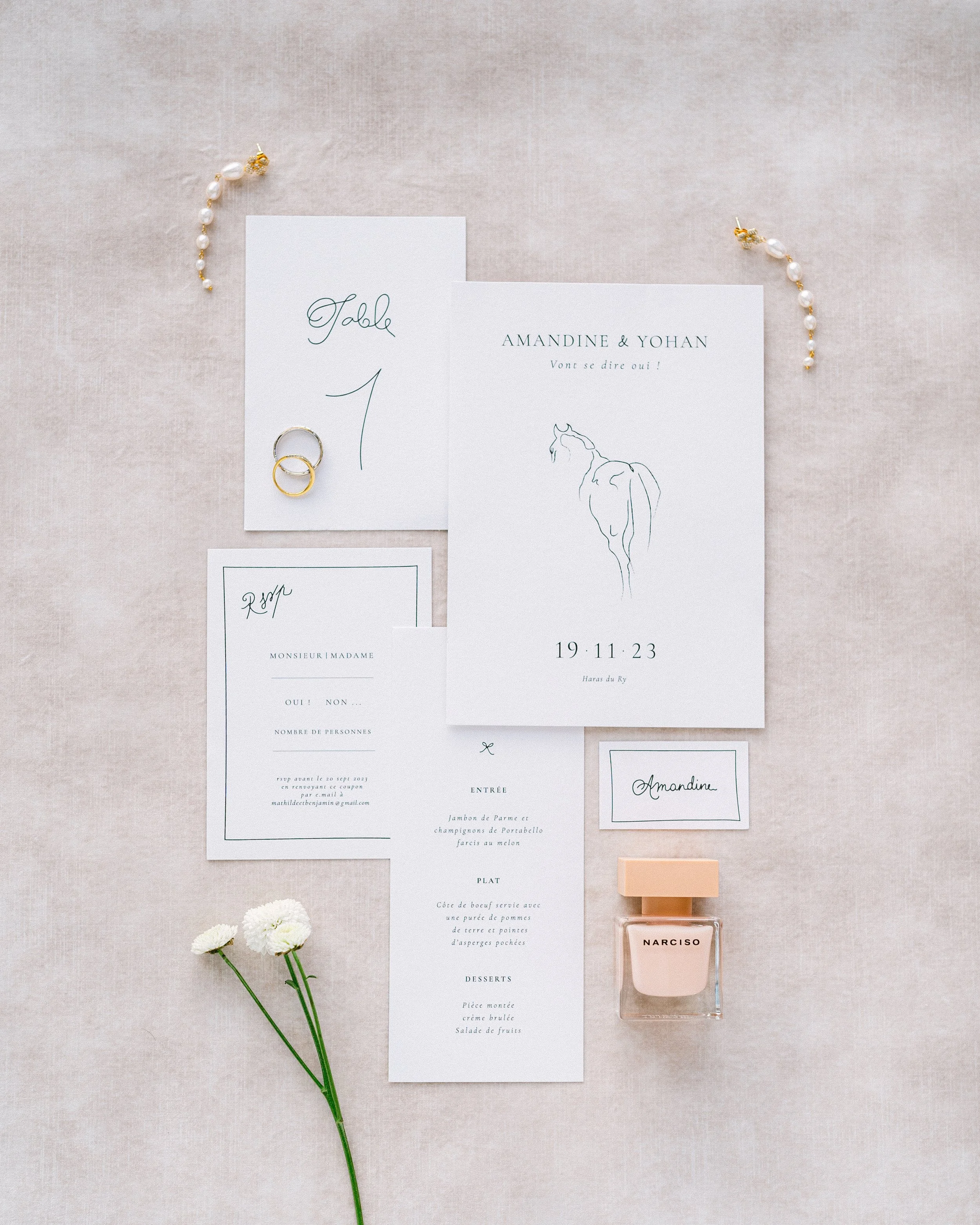 Invitation de mariage avec des papiers, des alliances, une bouteille de parfum, des bijoux, et des fleurs sur un fond en lin beige.