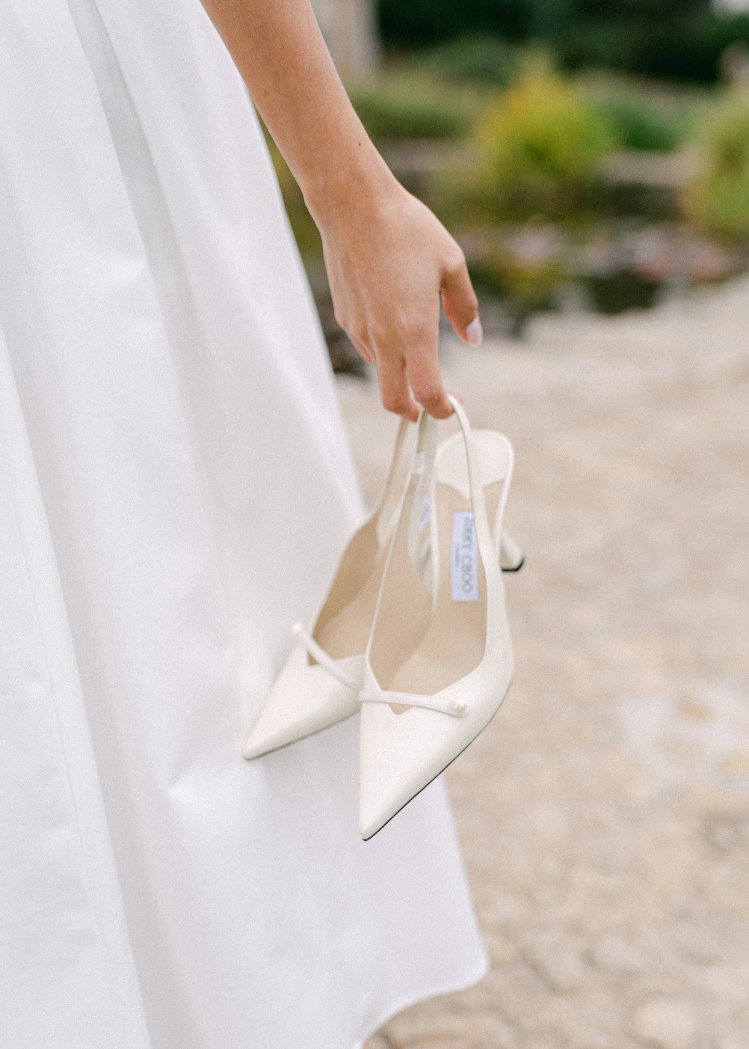 Une main tenant une paire de chaussures blanches à talons aiguilles, portée par une personne en robe blanche.