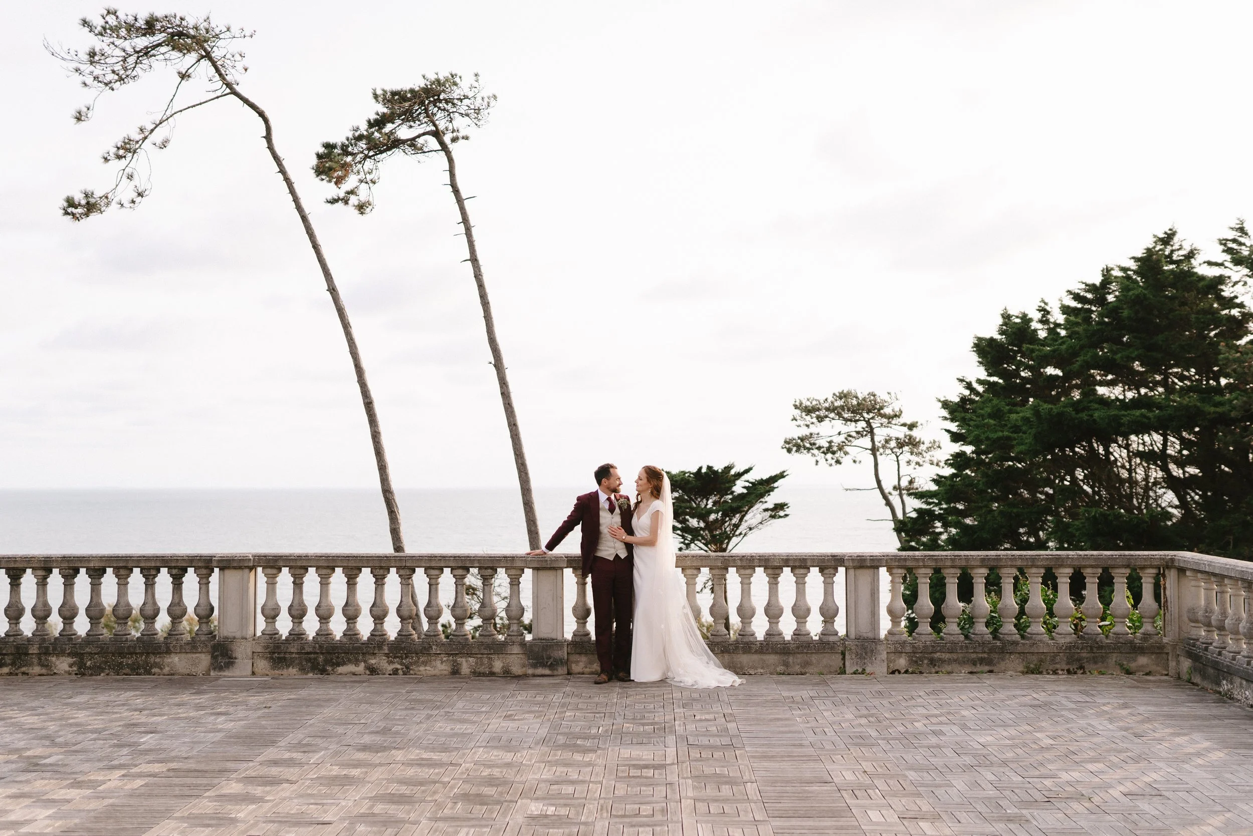 Un couple de mariés debout sur une terrasse en pierre avec une vue sur la mer, entourés d'arbres, lors d'un mariage