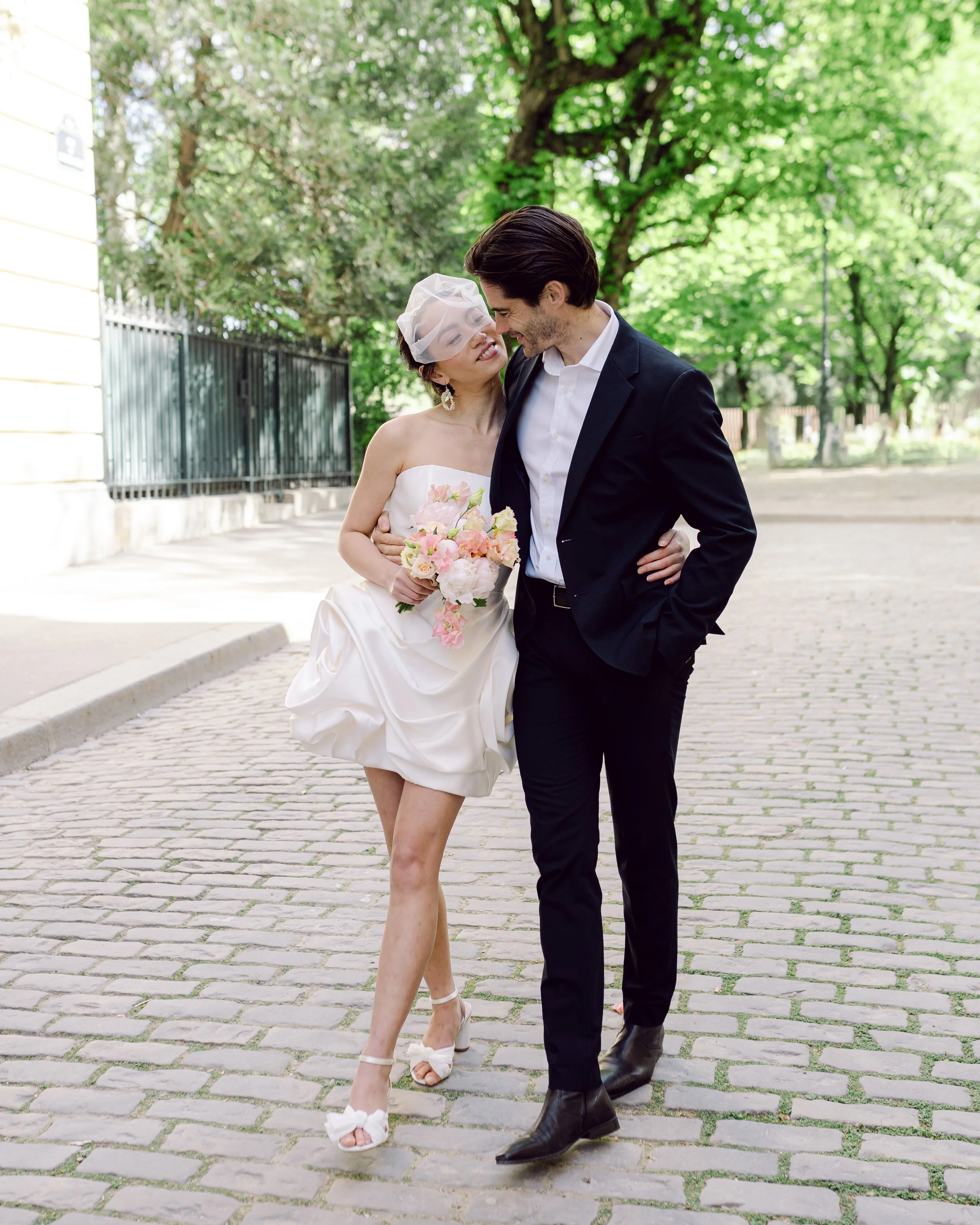 Un couple de mariés marche main dans la main en plein air, avec des arbres verts en arrière-plan.