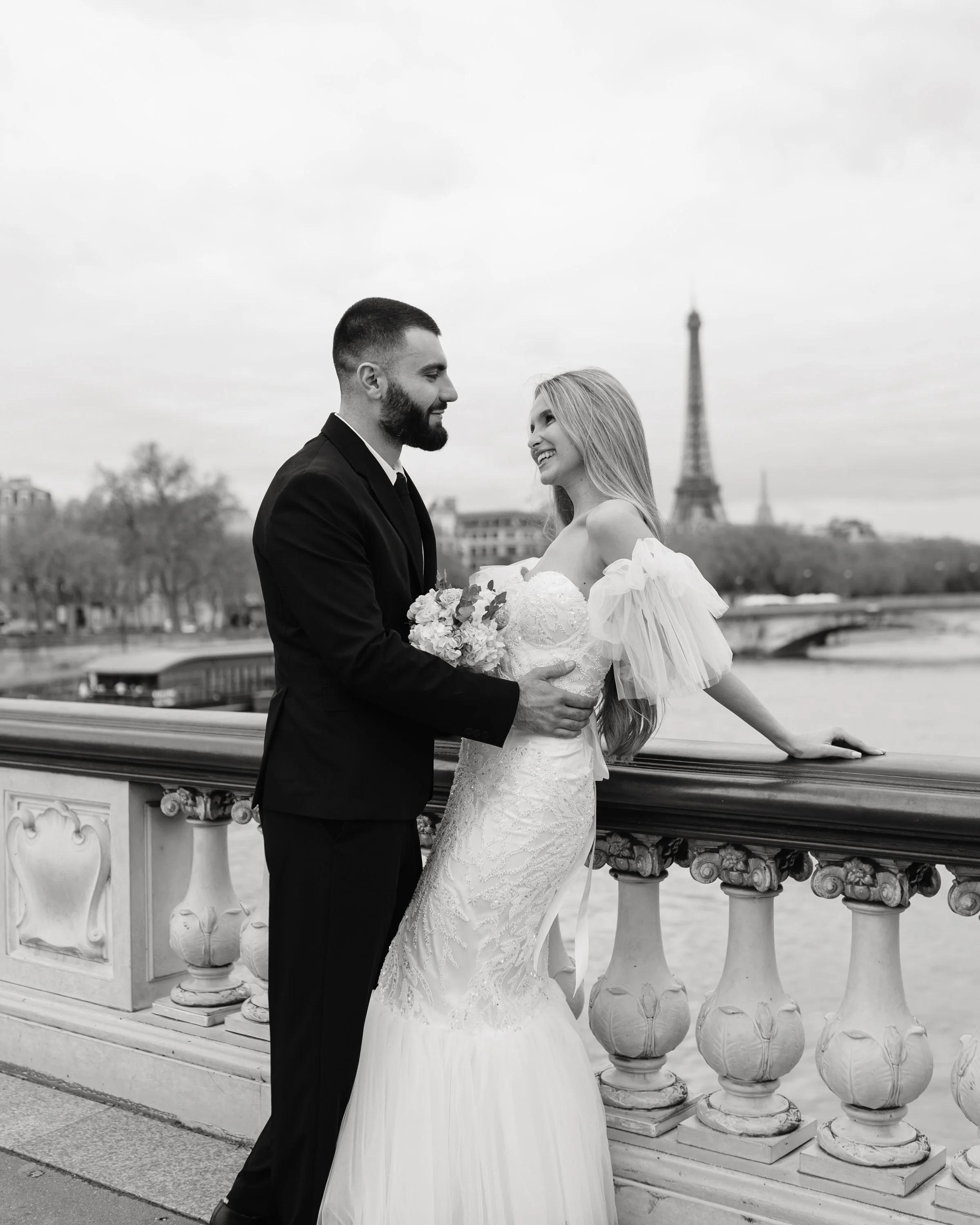 Un couple en mariage devant la Seine à Paris, avec la Tour Eiffel en arrière-plan, en noir et blanc.