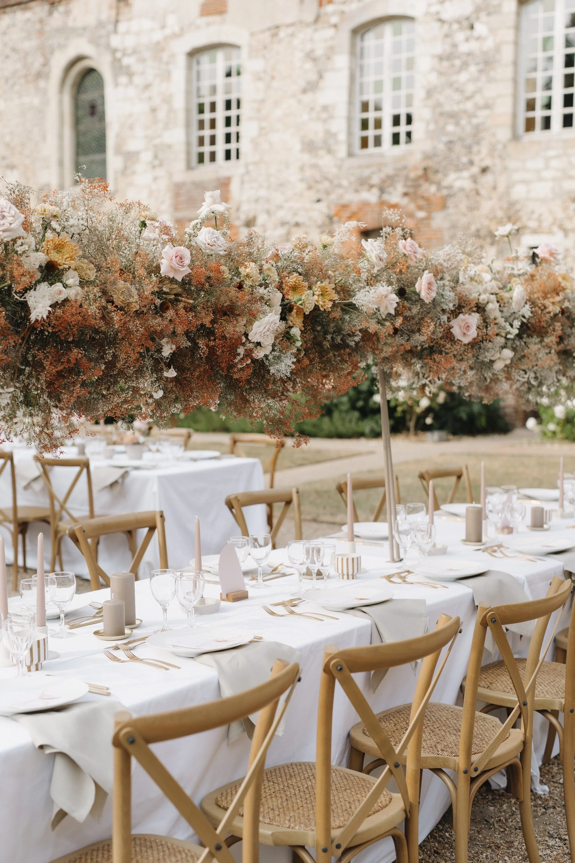 Table de mariage décorée avec une nappe blanche, des vaisselles, des verres à vin, des bougies et un grand centre de fleurs suspendu dans une cour extérieure avec un bâtiment en pierre en arrière-plan.
