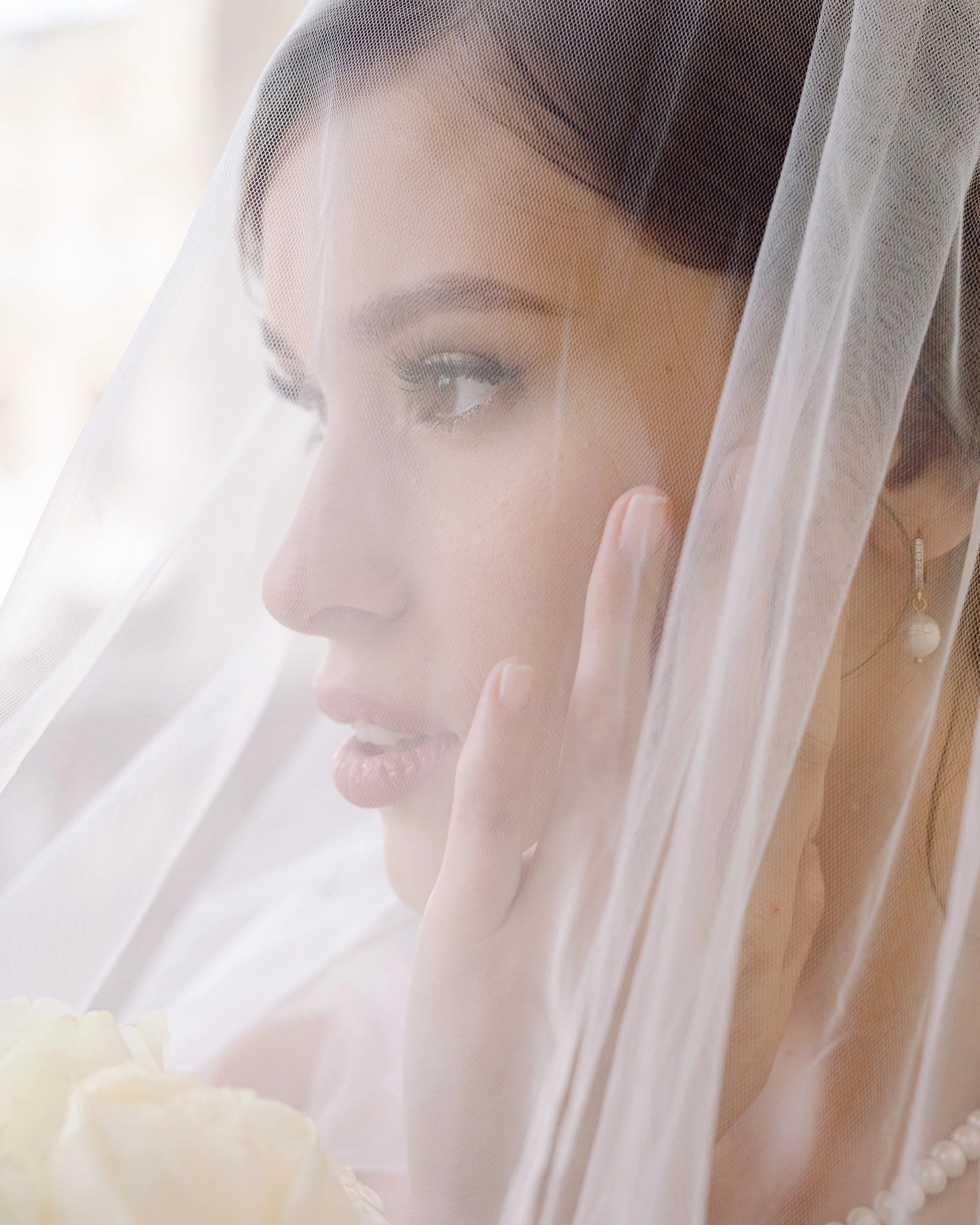 Femme portant un voile de mariée, regardant pensivement par une fenêtre, avec boucles d'oreilles perles.