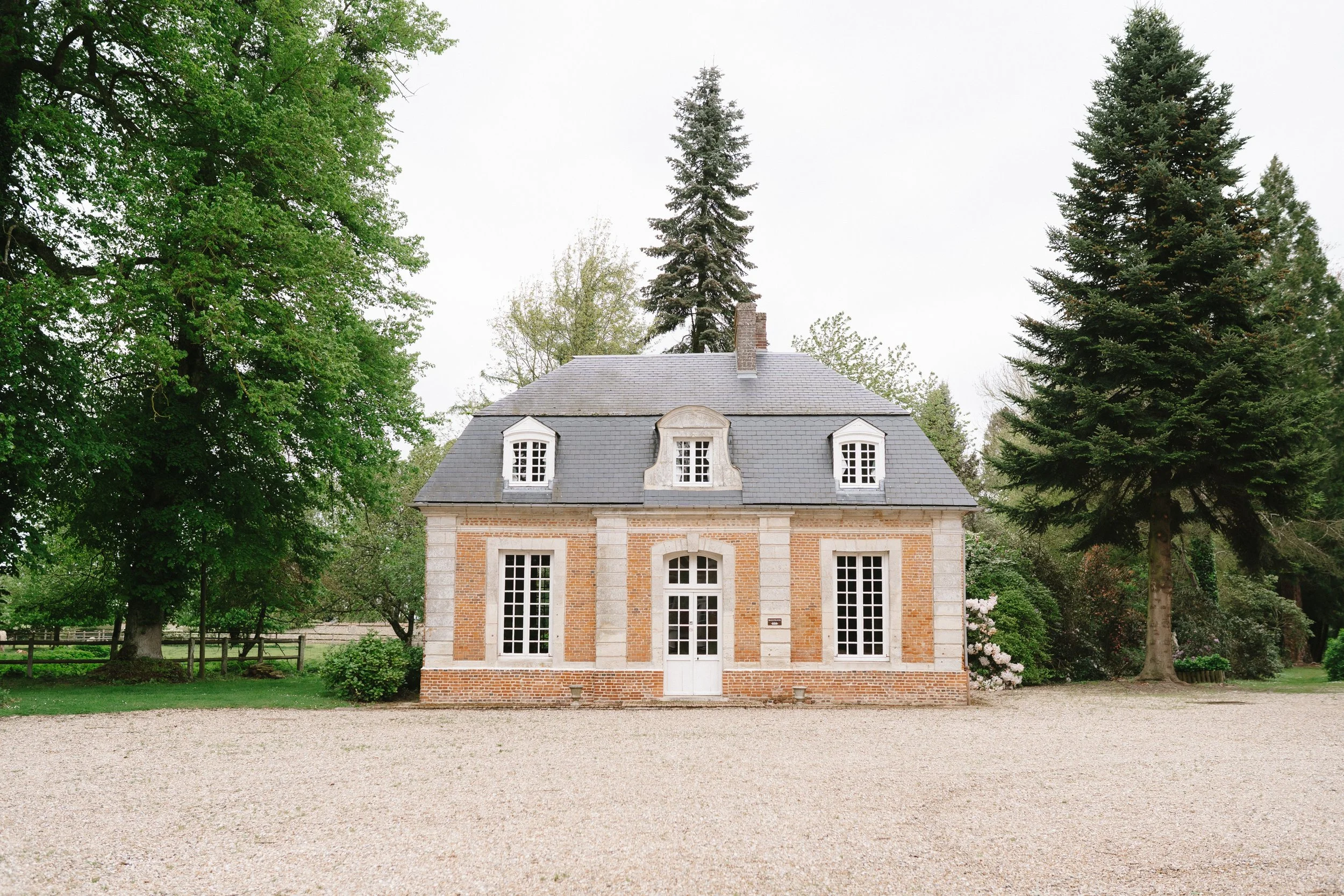 Une maison de style traditionnel en briques avec un toit en ardoise, entourée d'arbres verts, avec une allée en gravier devant.