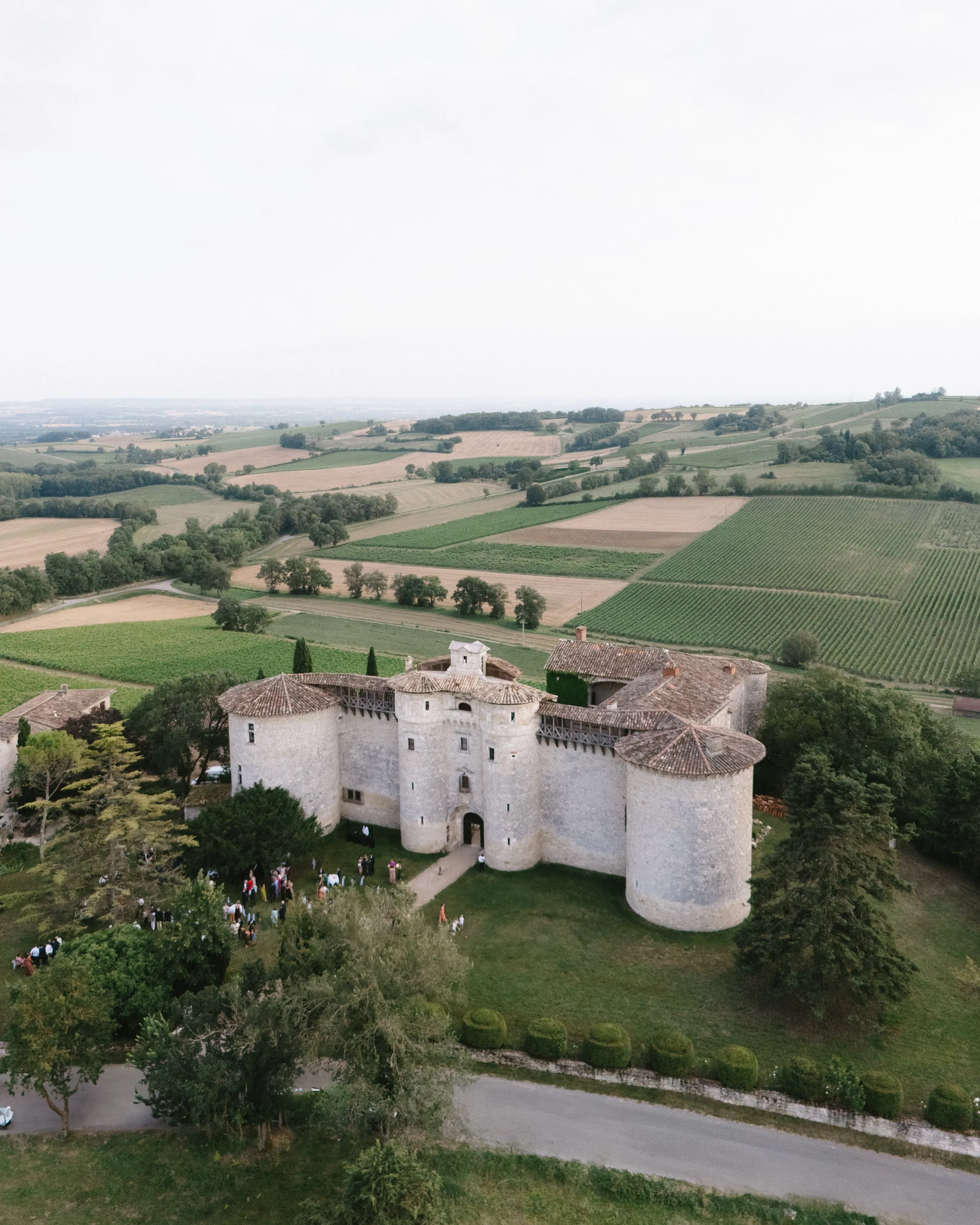 Château médiéval en pierre blanche avec tours rondes, entouré de verdure et de champs agricoles, avec un groupe de personnes à l'entrée, sous un ciel légèrement nuageux.