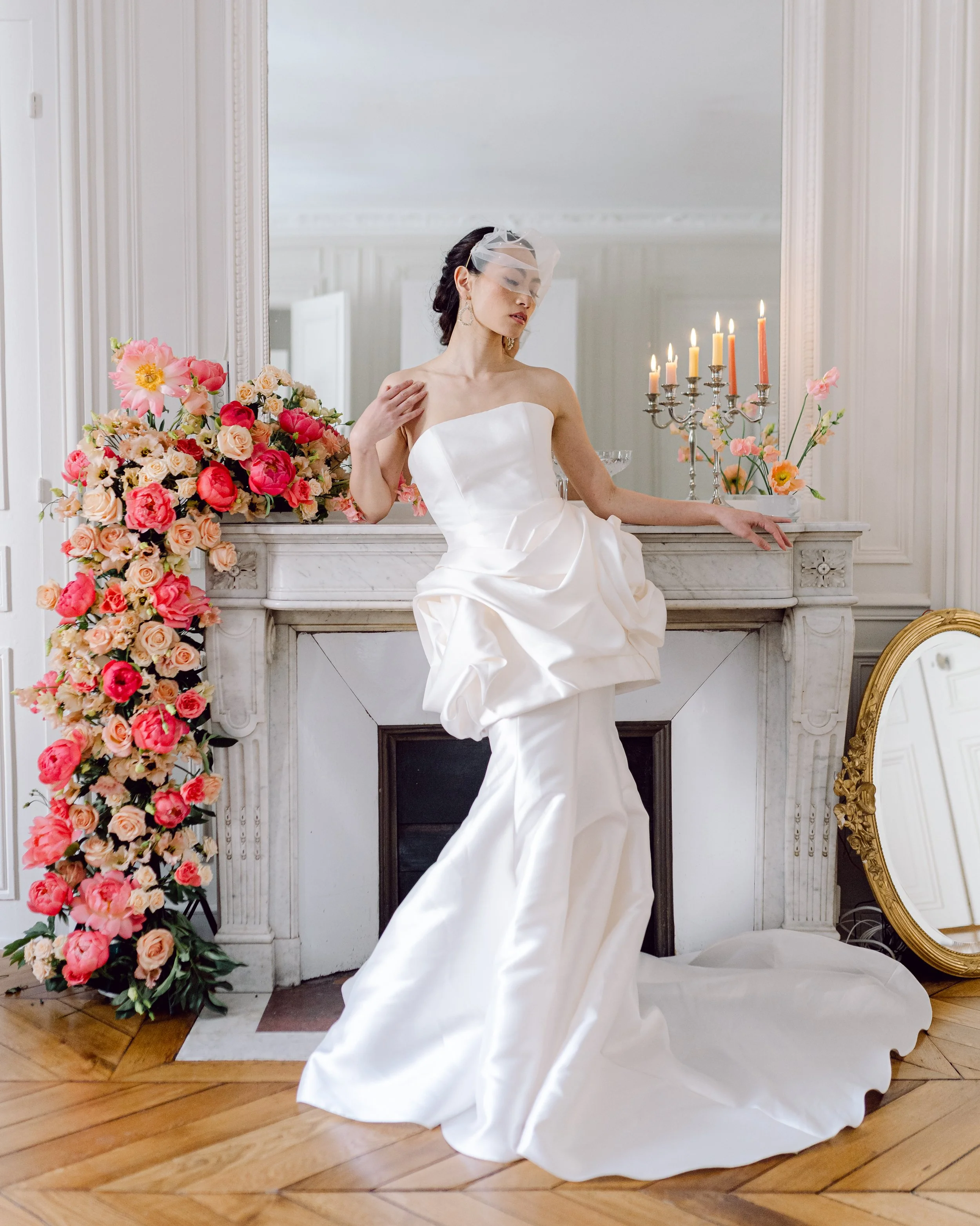 Une femme en robe de mariée blanche posant devant une cheminée décorée de fleurs et de chandelles, dans une pièce élégante avec un miroir à cadre doré à terre.