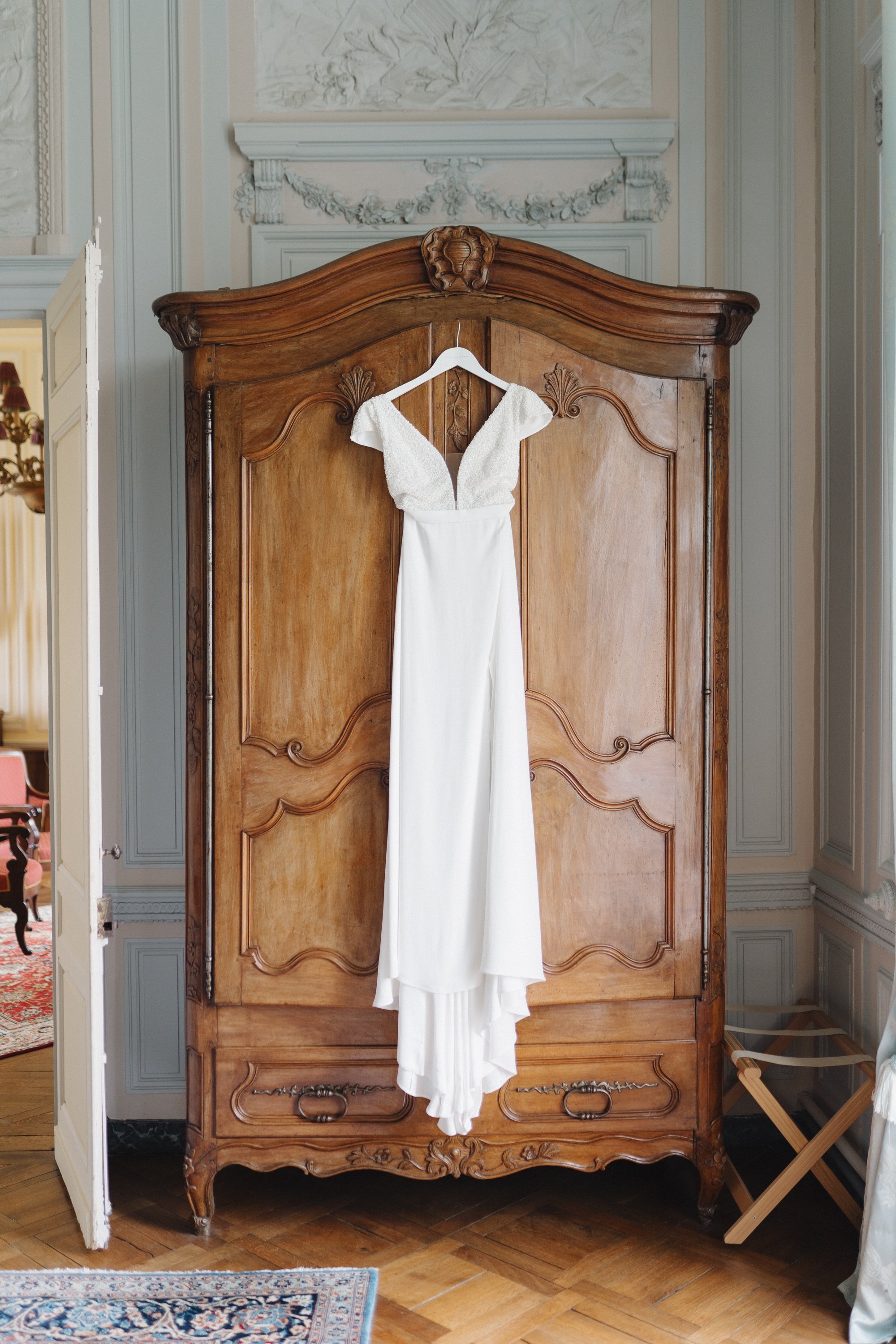 Une robe de mariée blanche suspendue à un meuble ancien en bois dans une pièce élégante avec des murs décorés.