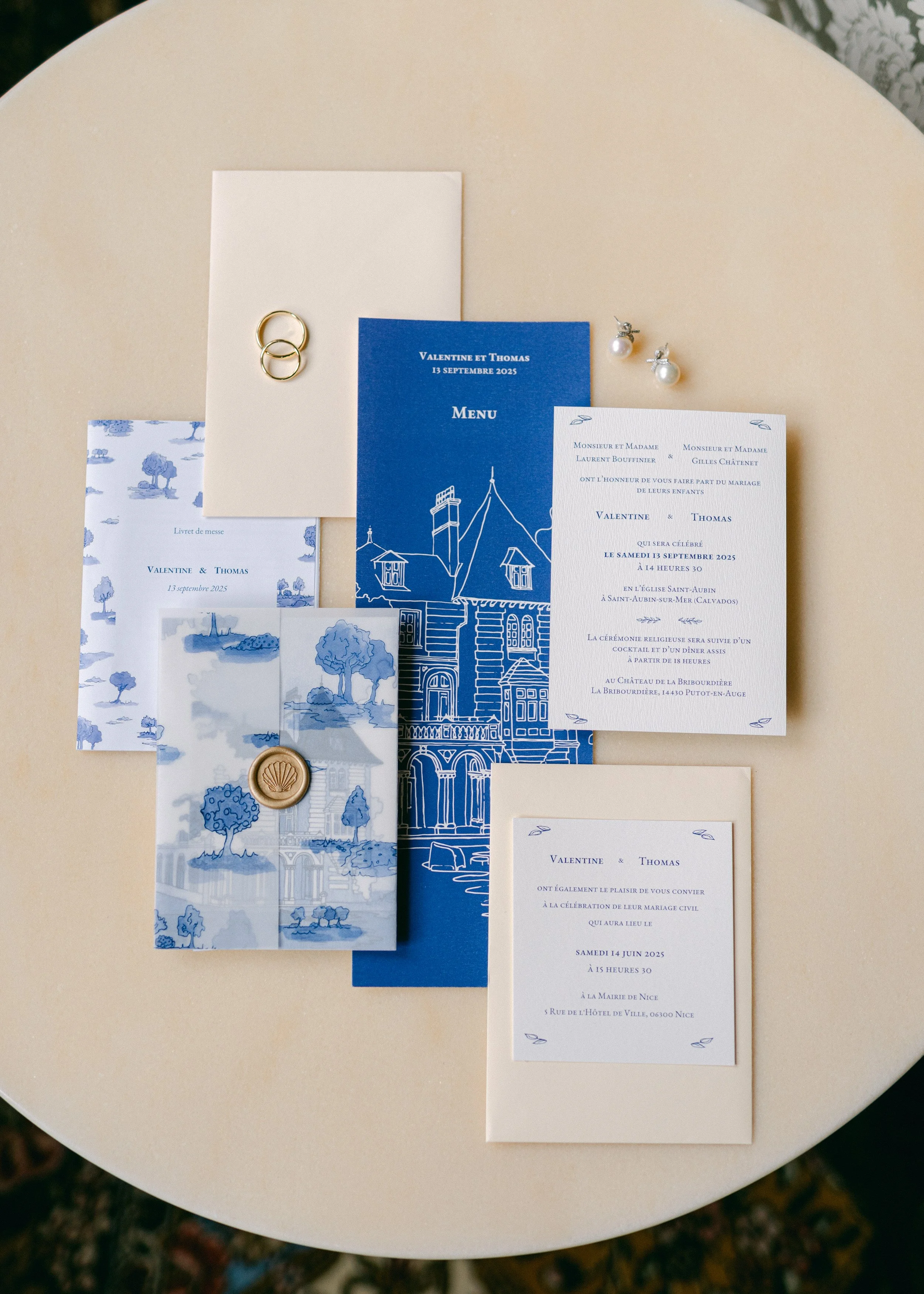 Invitation de mariage bleu et blanc avec accessoires comme des bagues, des boucles d'oreilles, et des cartes de vœux, disposés sur une table ronde de couleur claire.