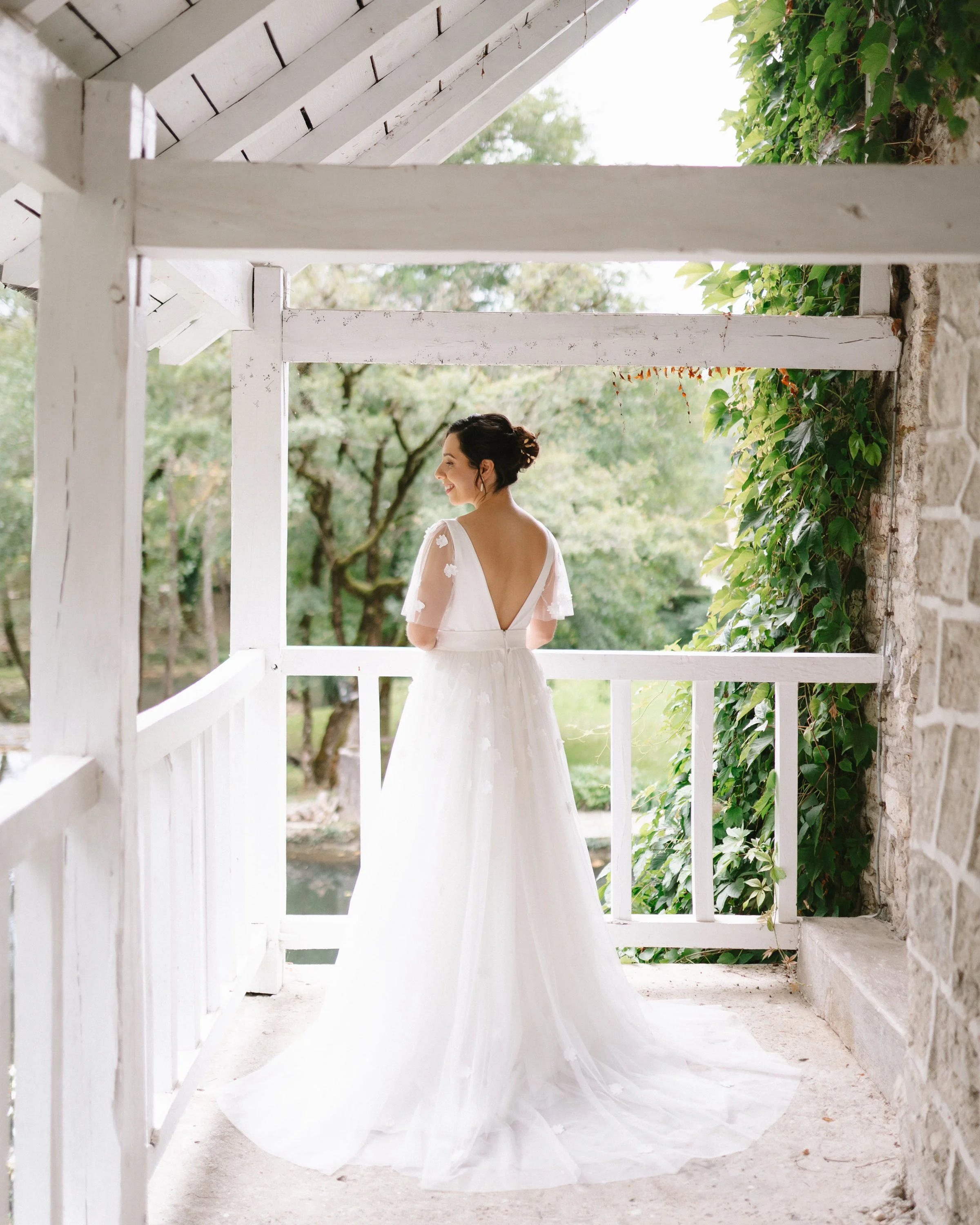 Une femme en robe de mariée blanche qui se tient sur un balcon en bois blanc, entourée de verdure.