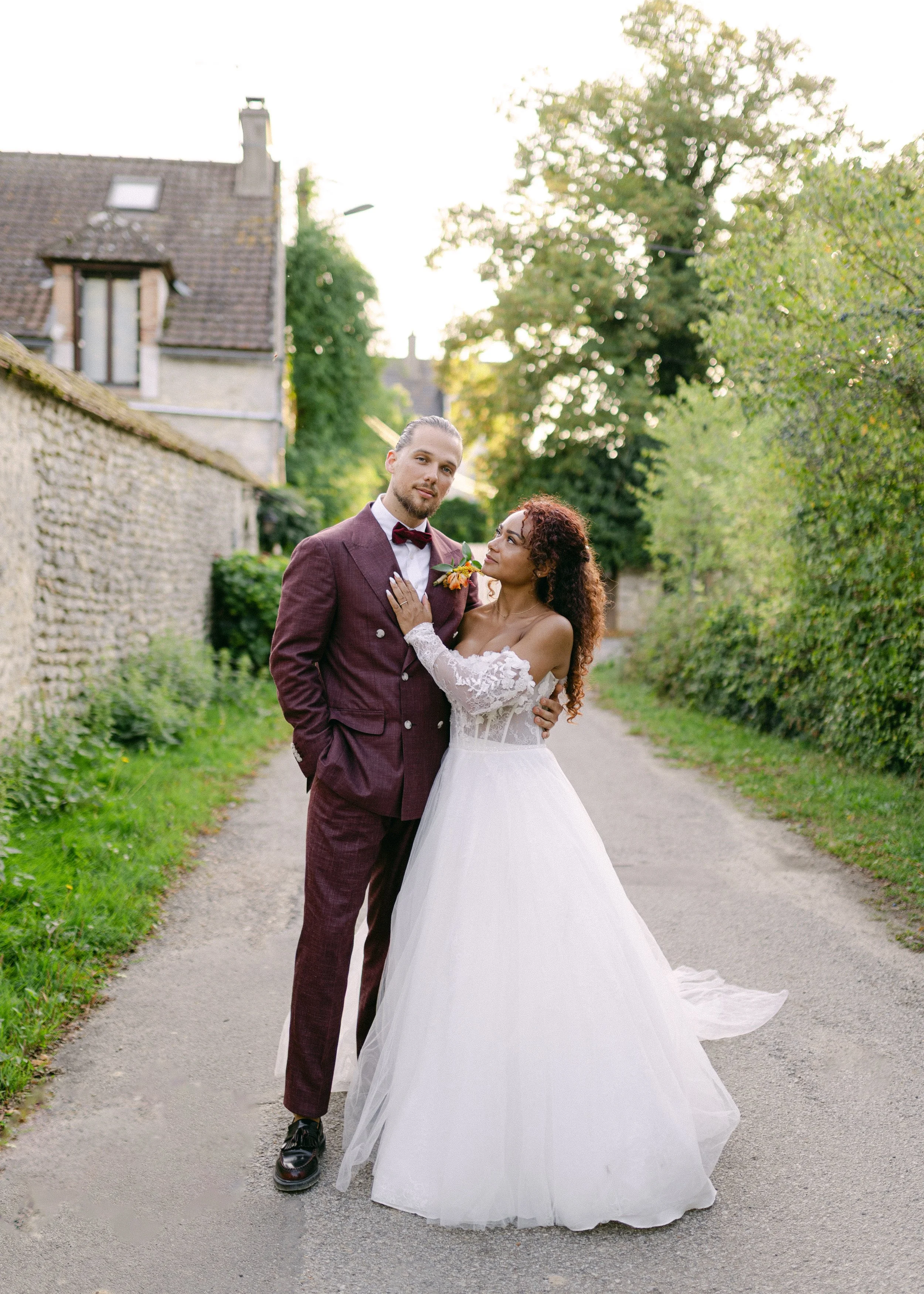 Un couple en costume de mariage posant dans une rue pavée entourée de verdure, le soleil se couchant à l'arrière-plan.