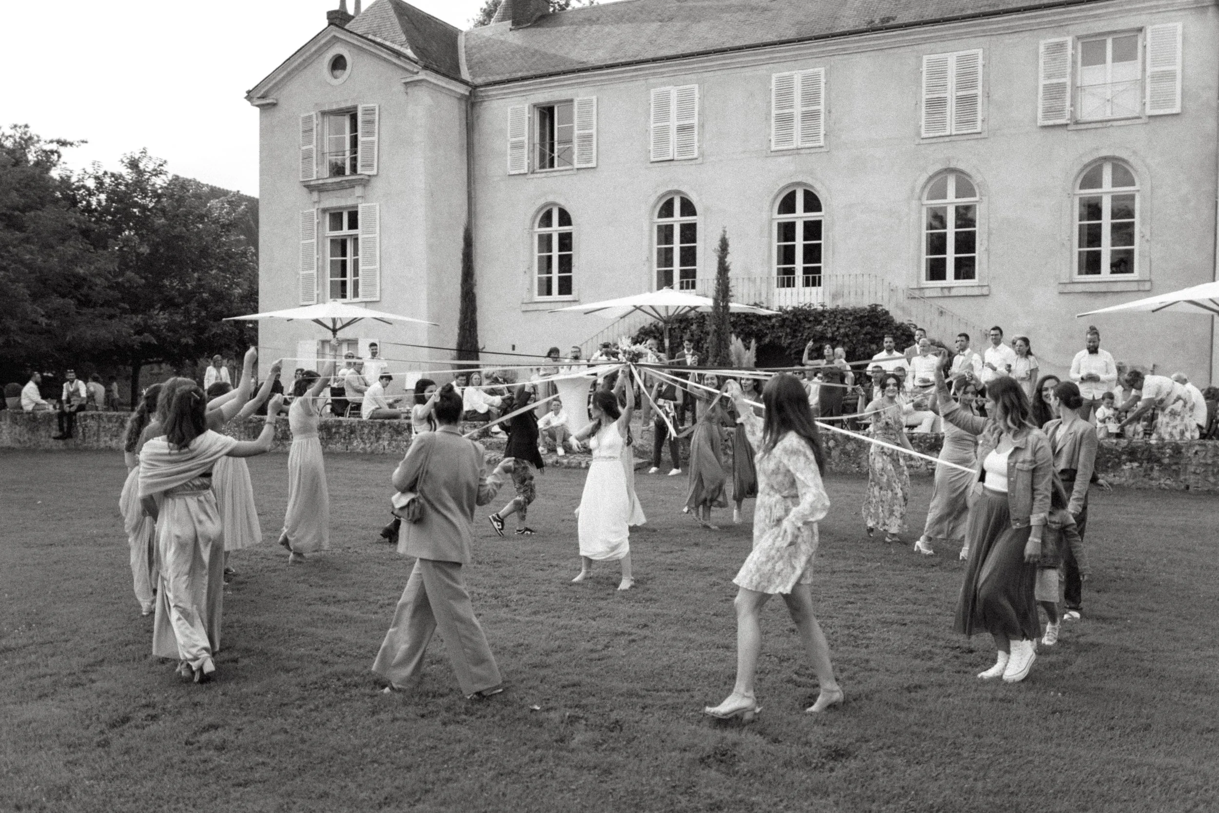Un groupe de personnes danse en cercle lors d'une fête en plein air devant une grande maison, avec des parasols et des spectateurs assis ou debout en arrière-plan.