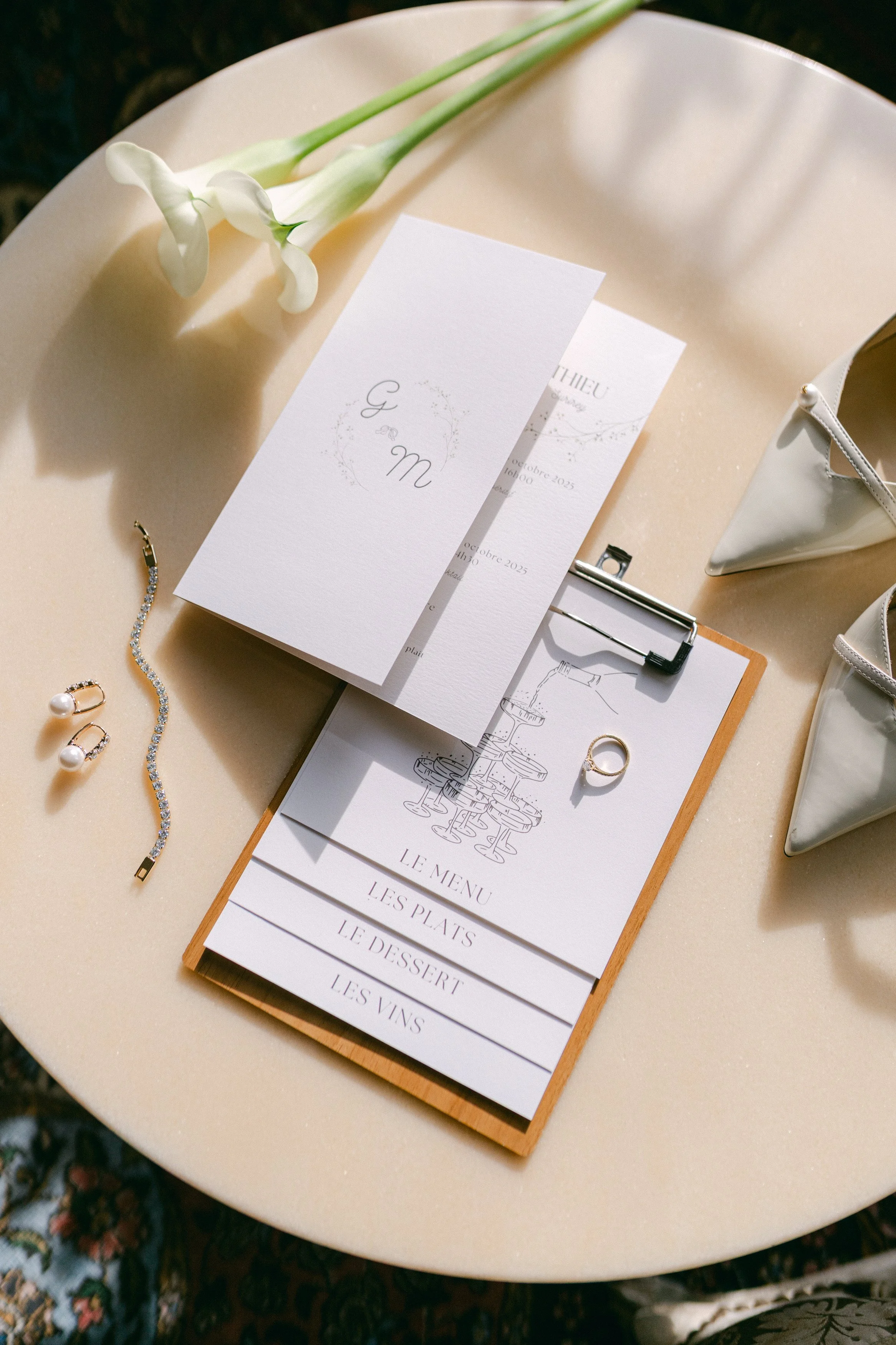 Une table ronde avec une invitation de mariage, un menu, des bijoux (boucles d'oreilles, bracelet), une bague, des fleurs blanches, et des sacs en tissu blanc.