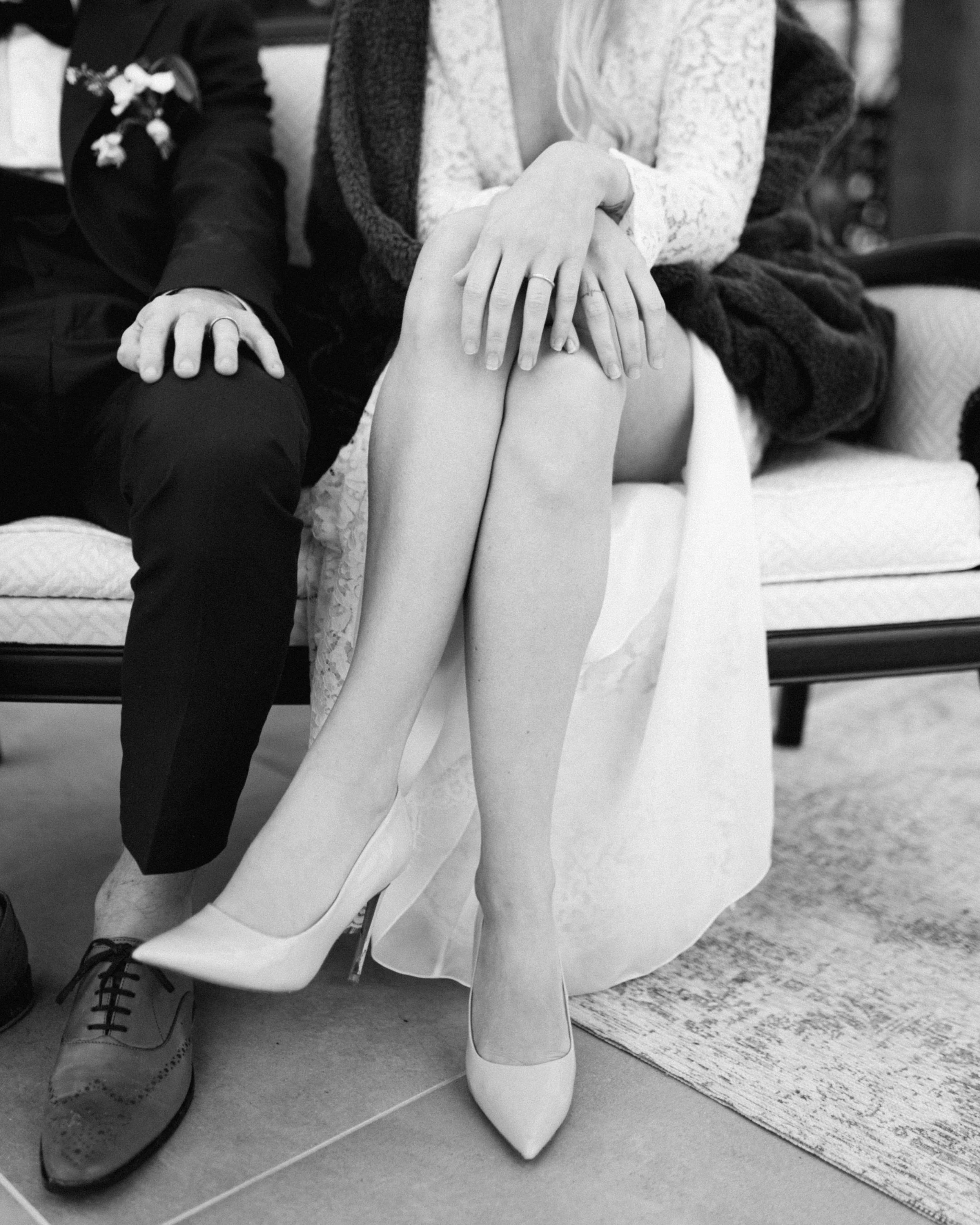 Une personne assise sur un canapé, portant une robe et des talons blancs, avec une main posée sur une autre personne vêtue d'un costume. La scène semble intime et élégante.