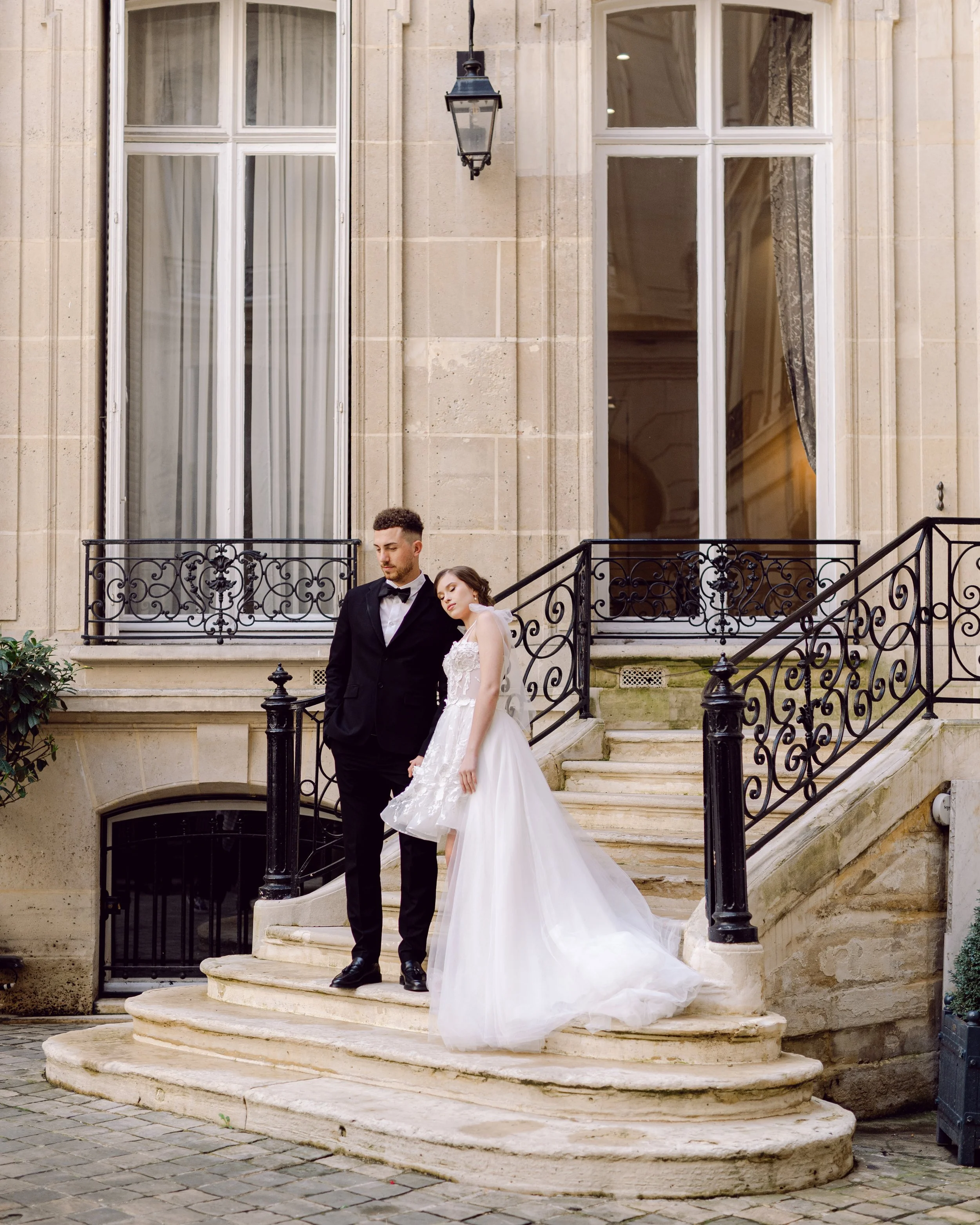 Un couple en mariage pose sur un escalier en pierre devant une grande maison en pierre avec de grandes fenêtres. La mariée porte une robe blanche et le marié un costume noir avec une chemise blanche