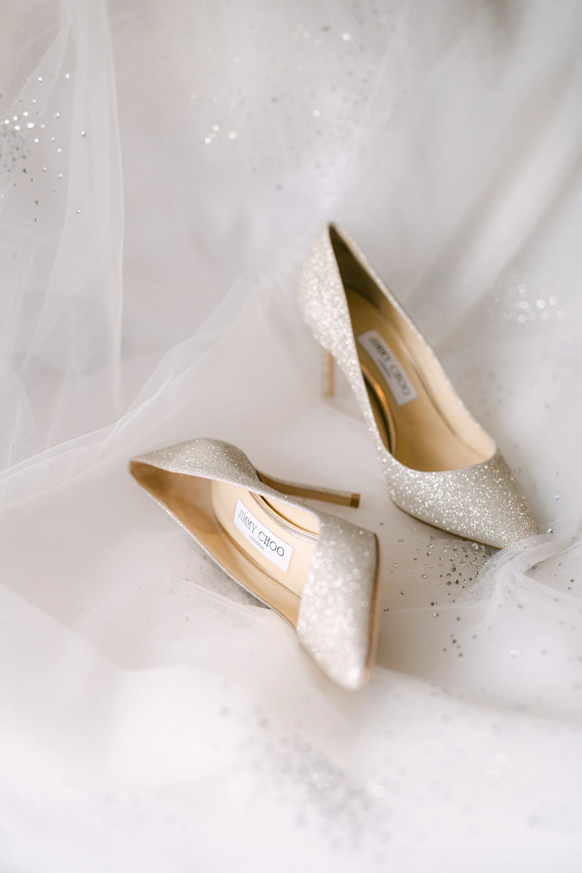 Une paire de chaussures à talons sandales en glitter argenté posée sur un tissu blanc avec des paillettes.