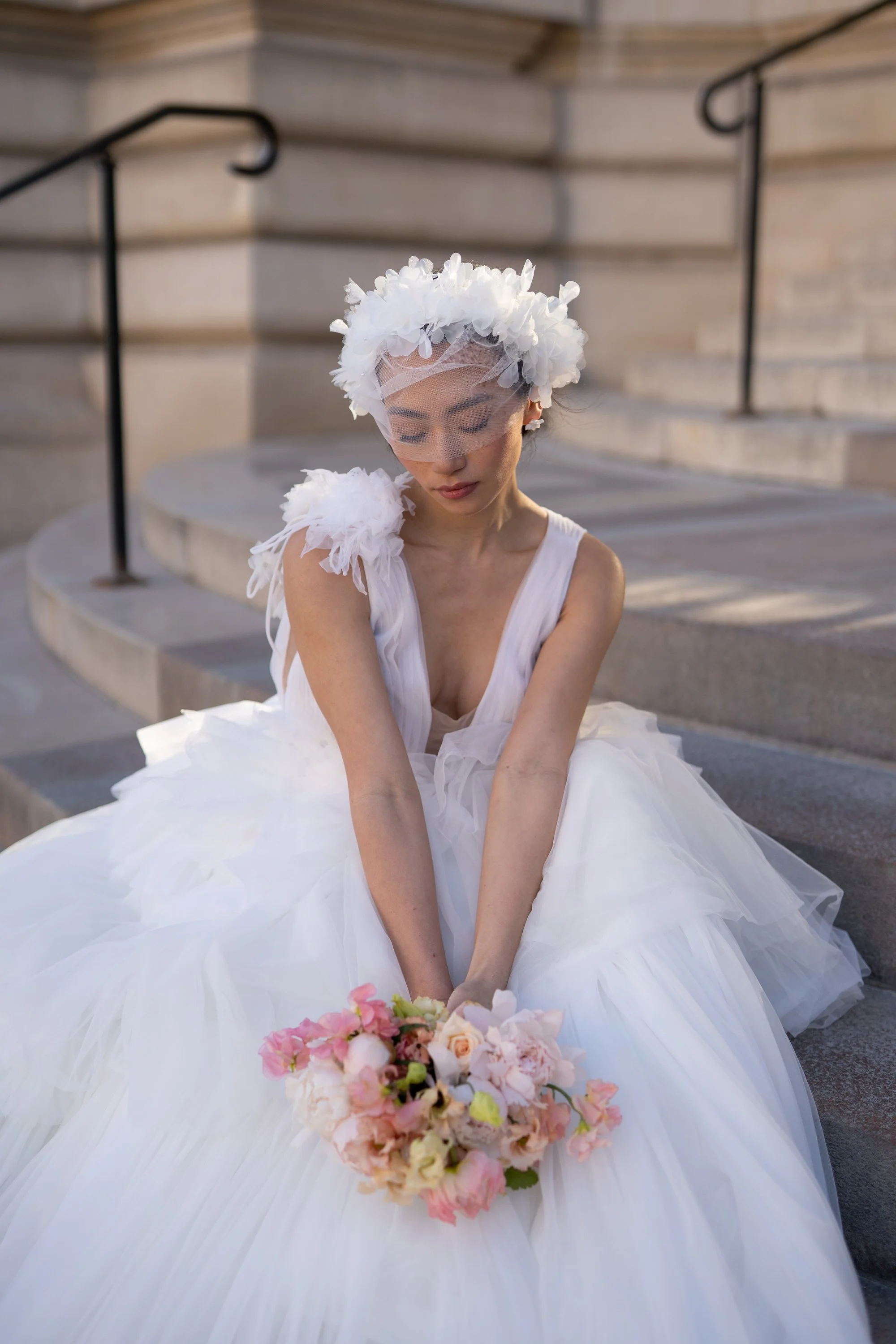 Une femme en robe de mariée blanche portant un voile et un collier de fleurs, assise sur des marches en pierre, tenant un bouquet de fleurs roses et blanches, avec une main reposant doucement sur ses genoux.