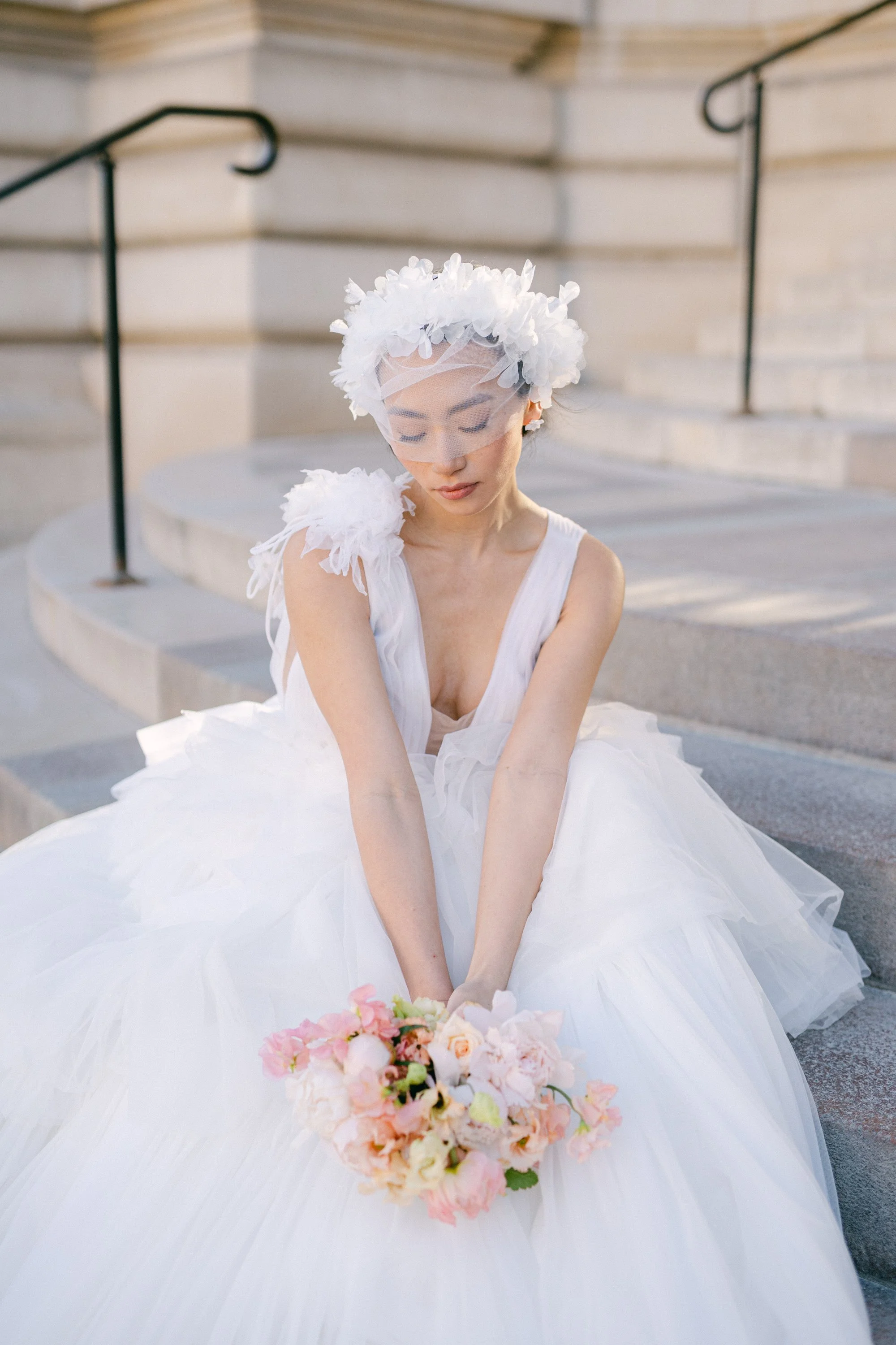 Femme en robe blanche de mariage assise sur des marches, portant un voile et un chapeau de fleurs blanches, tenant un bouquet de fleurs roses et blanches.