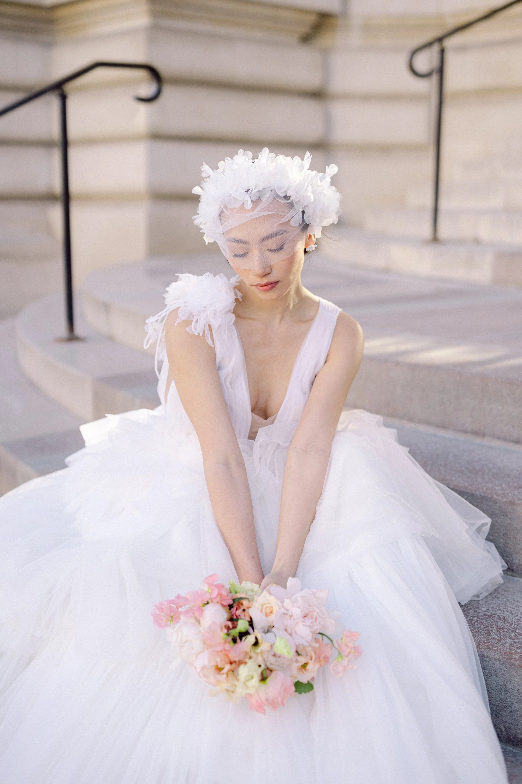 Jeune femme en robe blanche de mariage avec un voile et un bouquet de fleurs, assise sur des marches en pierre avec une rampe en métal.