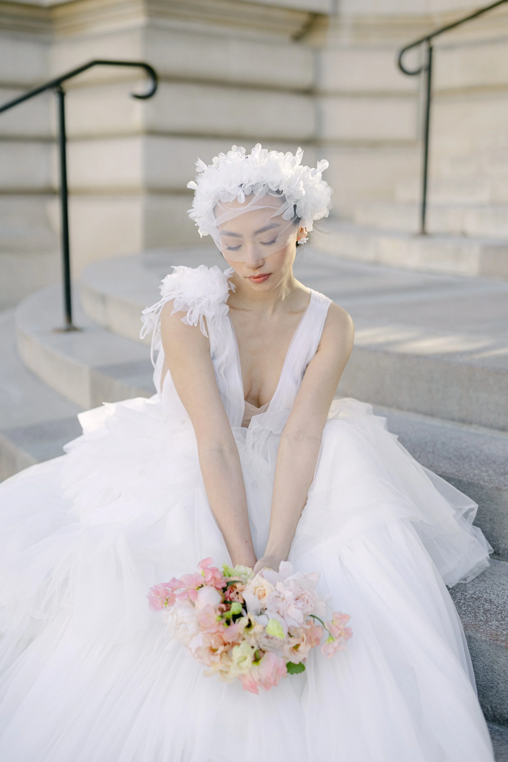 Jeune femme en robe de mariée blanche, portant un voile et un couronne de fleurs, assise sur des escaliers en pierre, tenant un bouquet de fleurs pastel.
