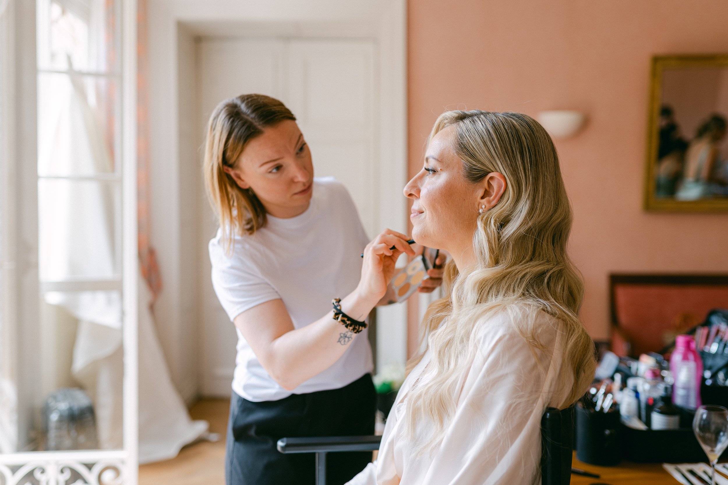 une femme fait du maquillage à une autre femme dans une pièce lumineuse avec des murs rose et une fenêtre