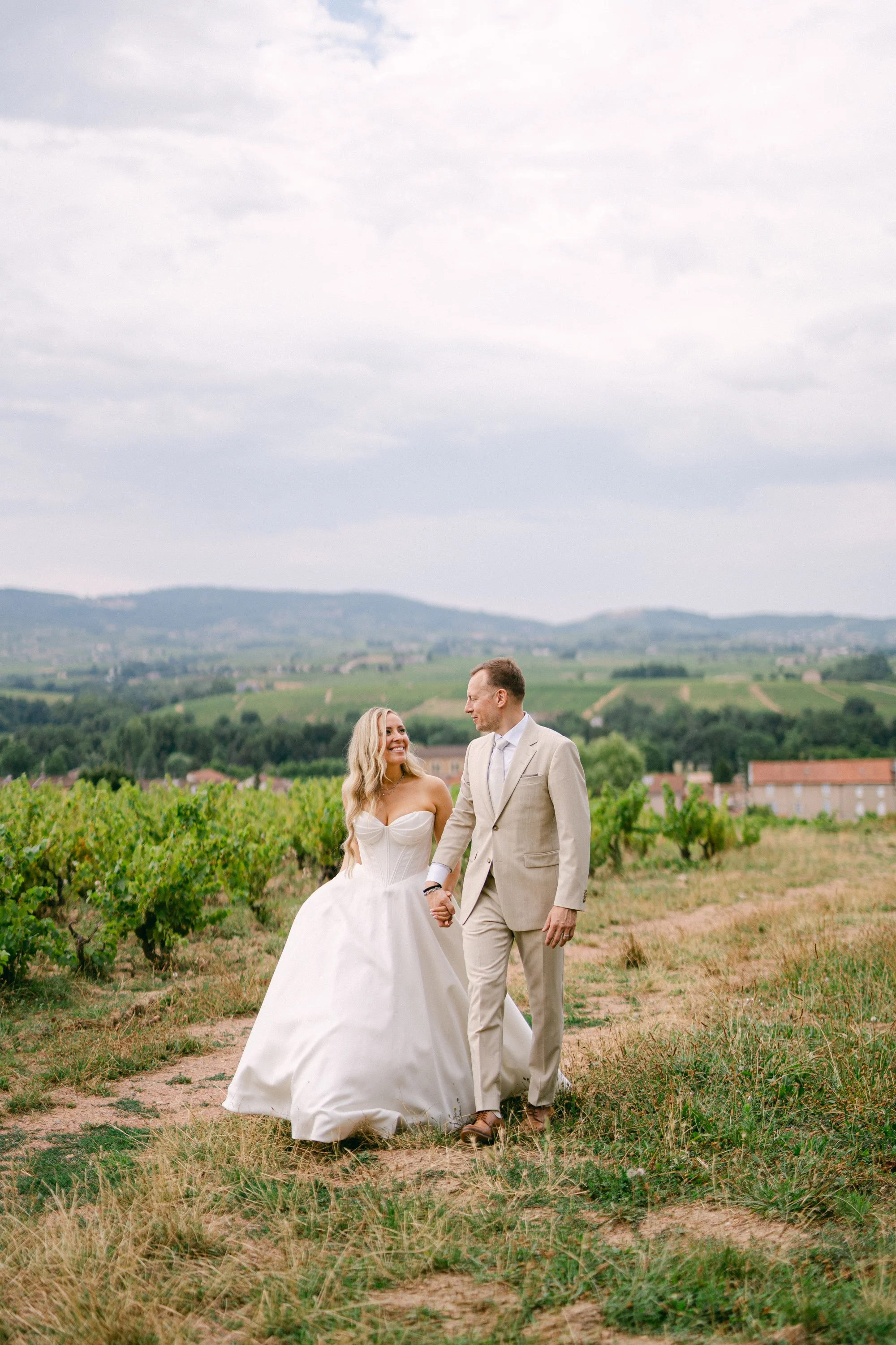 Un couple de mariés marche dans un champ avec des vignes, main dans la main, sous un ciel nuageux, avec un paysage de collines en arrière-plan.