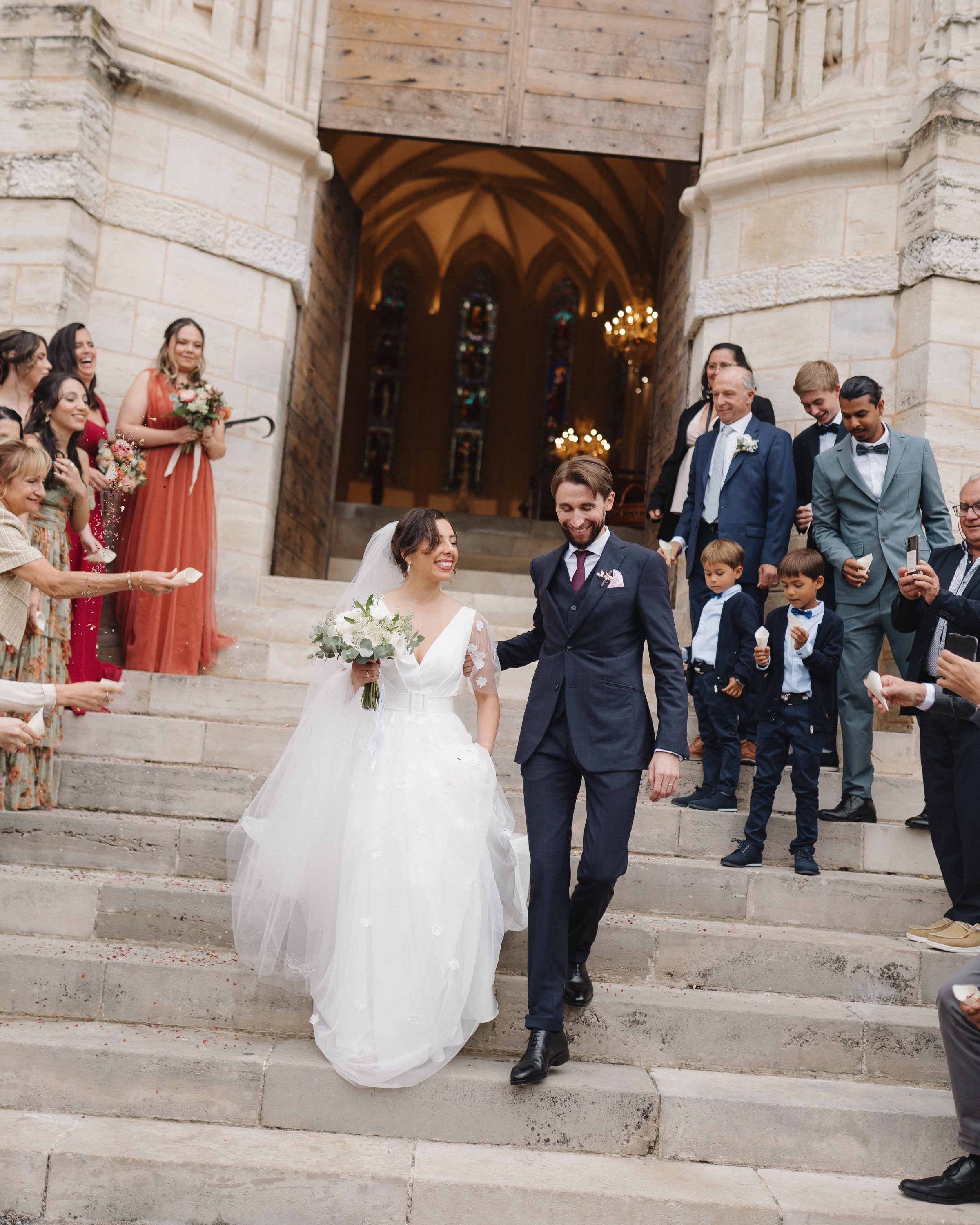 Un couple de mariés descend les escaliers d'une église, entouré de famille et amis célébrant leur mariage.
