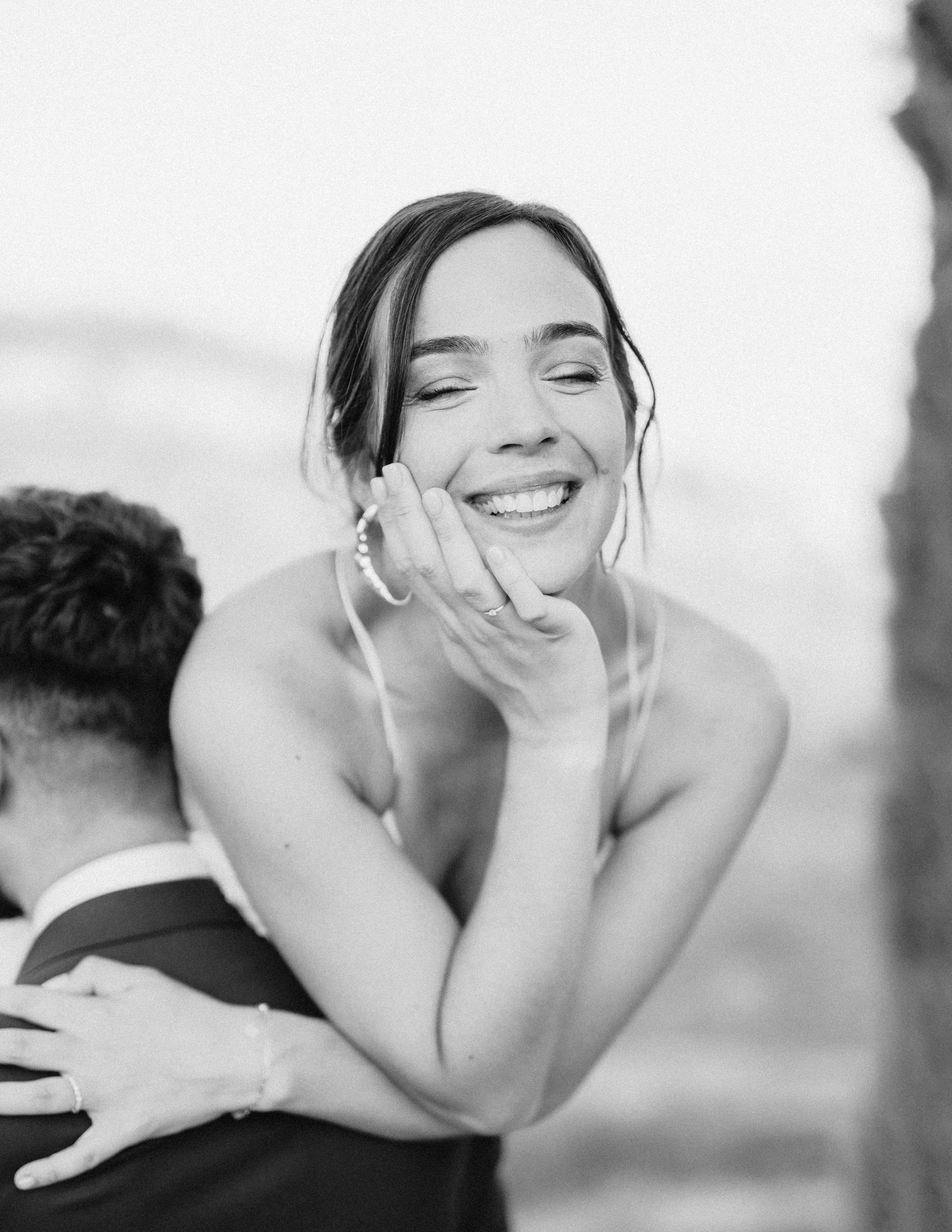 Une femme souriante en noir et blanc, tenant sa tête avec sa main, tandis qu'une personne dont la tête est coupée est en face d'elle. La femme porte un haut à baillettes et des boucles d'oreilles, avec une expression joyeuse.